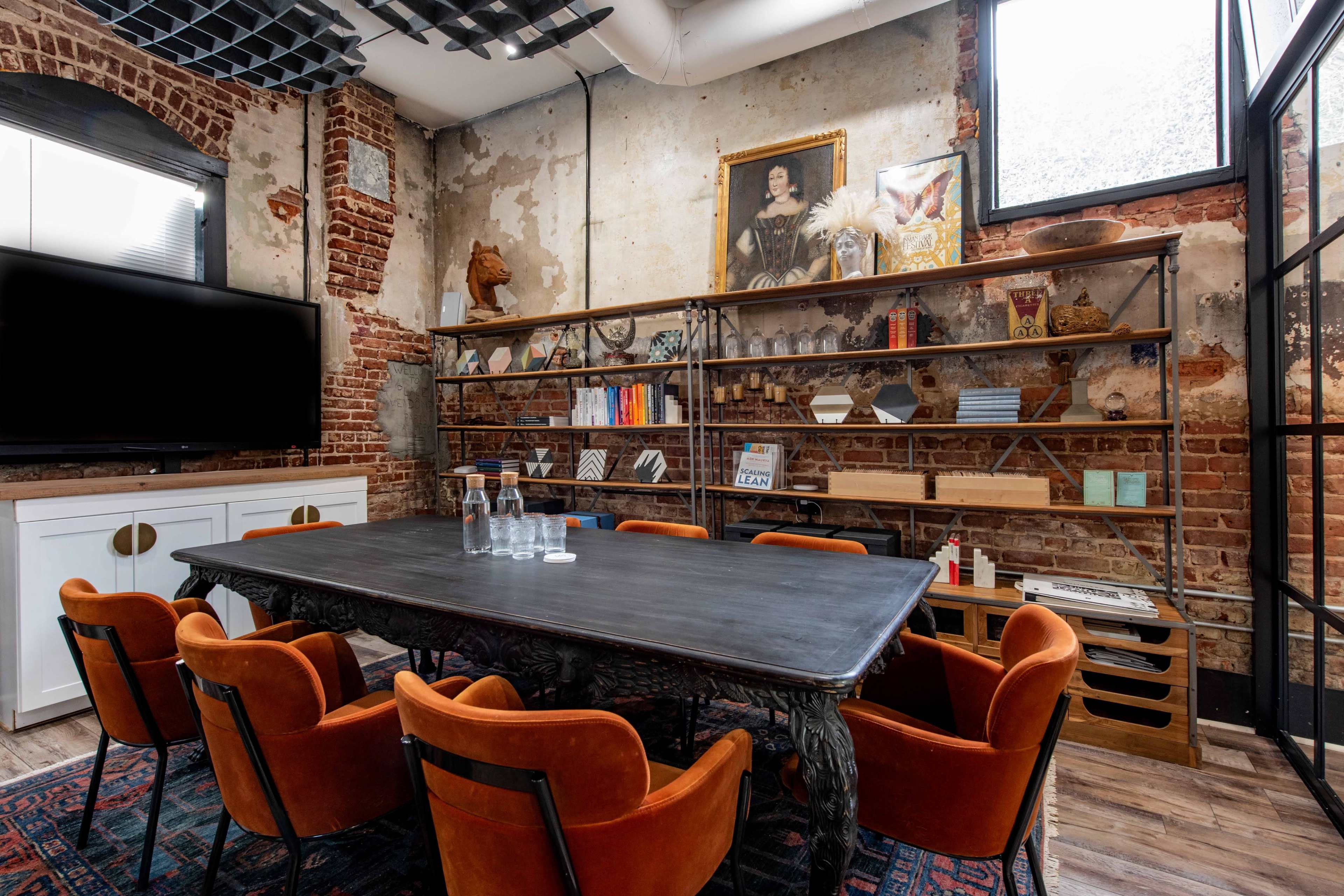 A conference room features a large dark table surrounded by orange chairs, with exposed brick walls and a variety of shelves displaying decor and books.