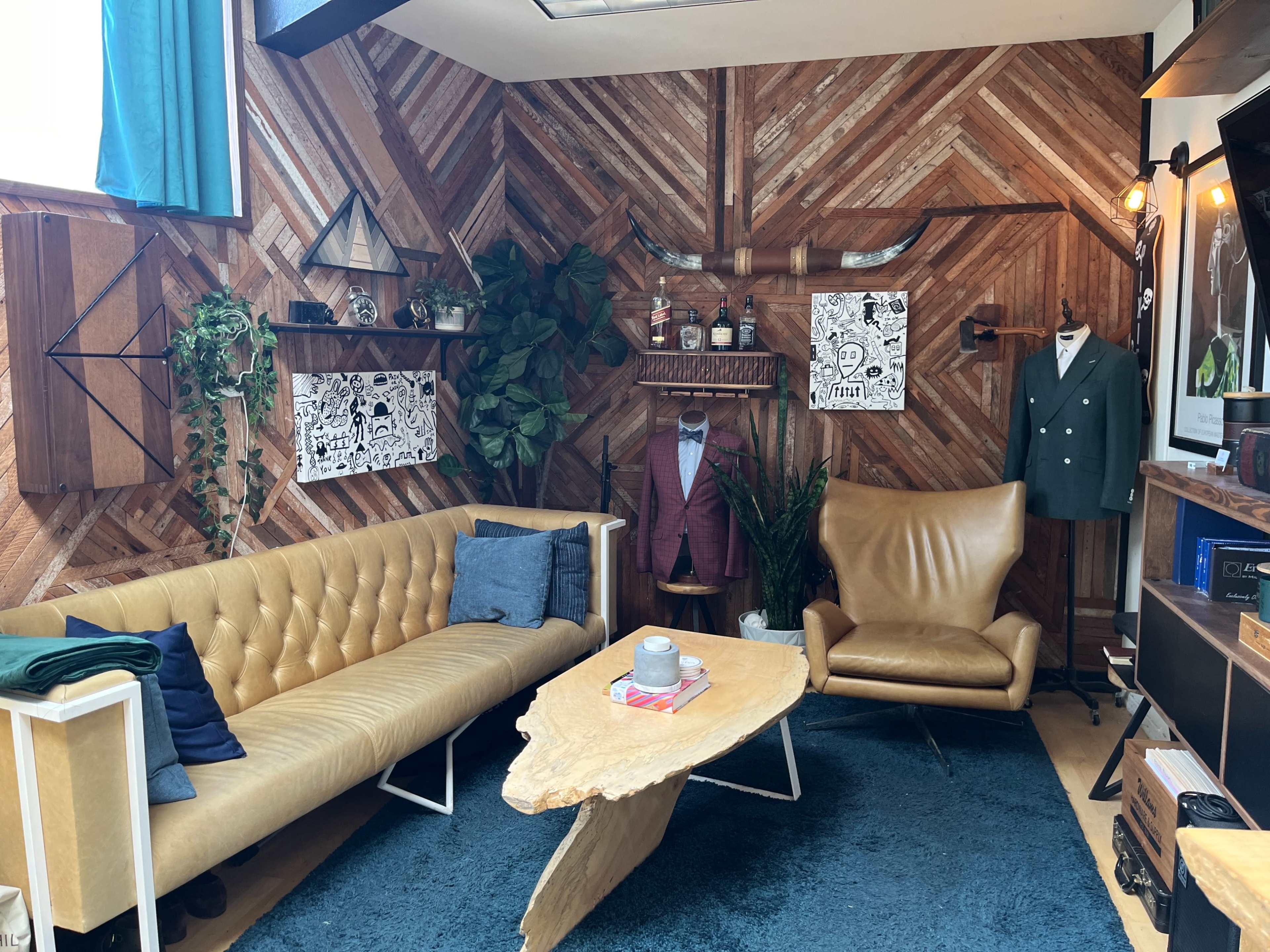 A stylish interior featuring a tan leather sofa, a wooden coffee table, and a green suit on display against a backdrop of patterned wood paneling and decorative plants.