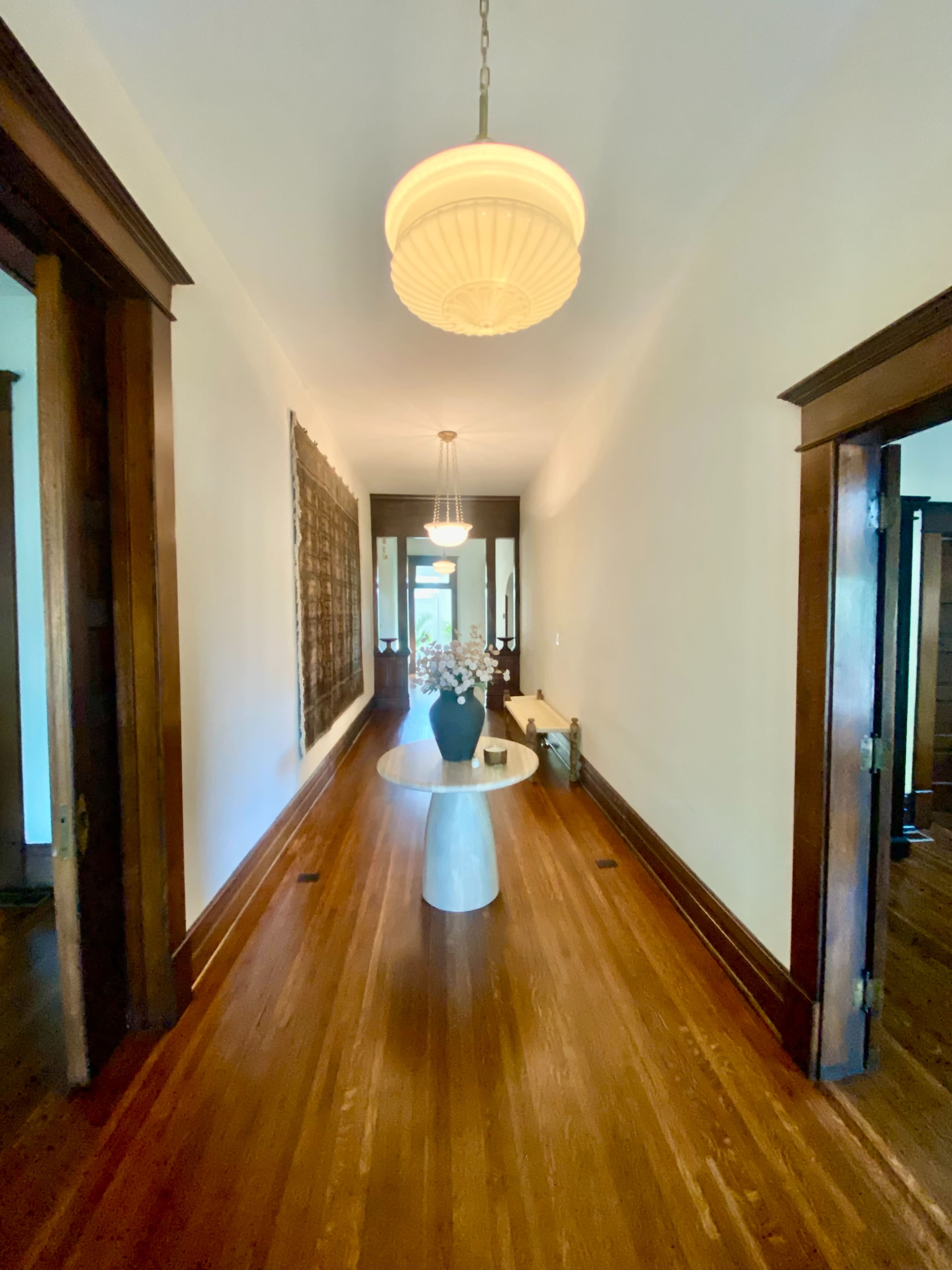 A long hallway features a round table with a vase of flowers, flanked by wooden doorways and illuminated by pendant lights.
