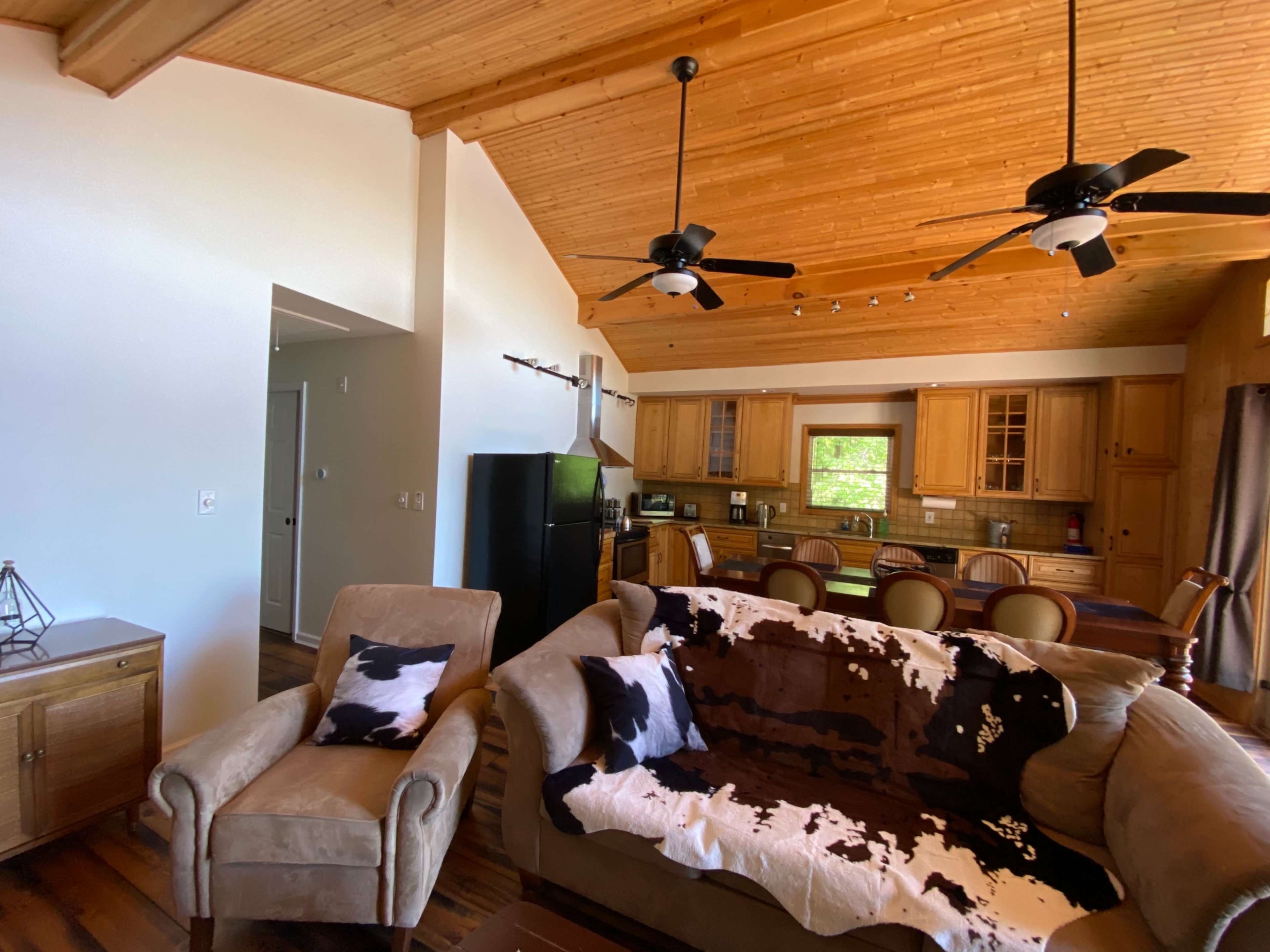 The image shows a spacious living room with a high wooden ceiling, featuring a sofa draped with a cowhide blanket, a kitchen area in the background, and a dining table.