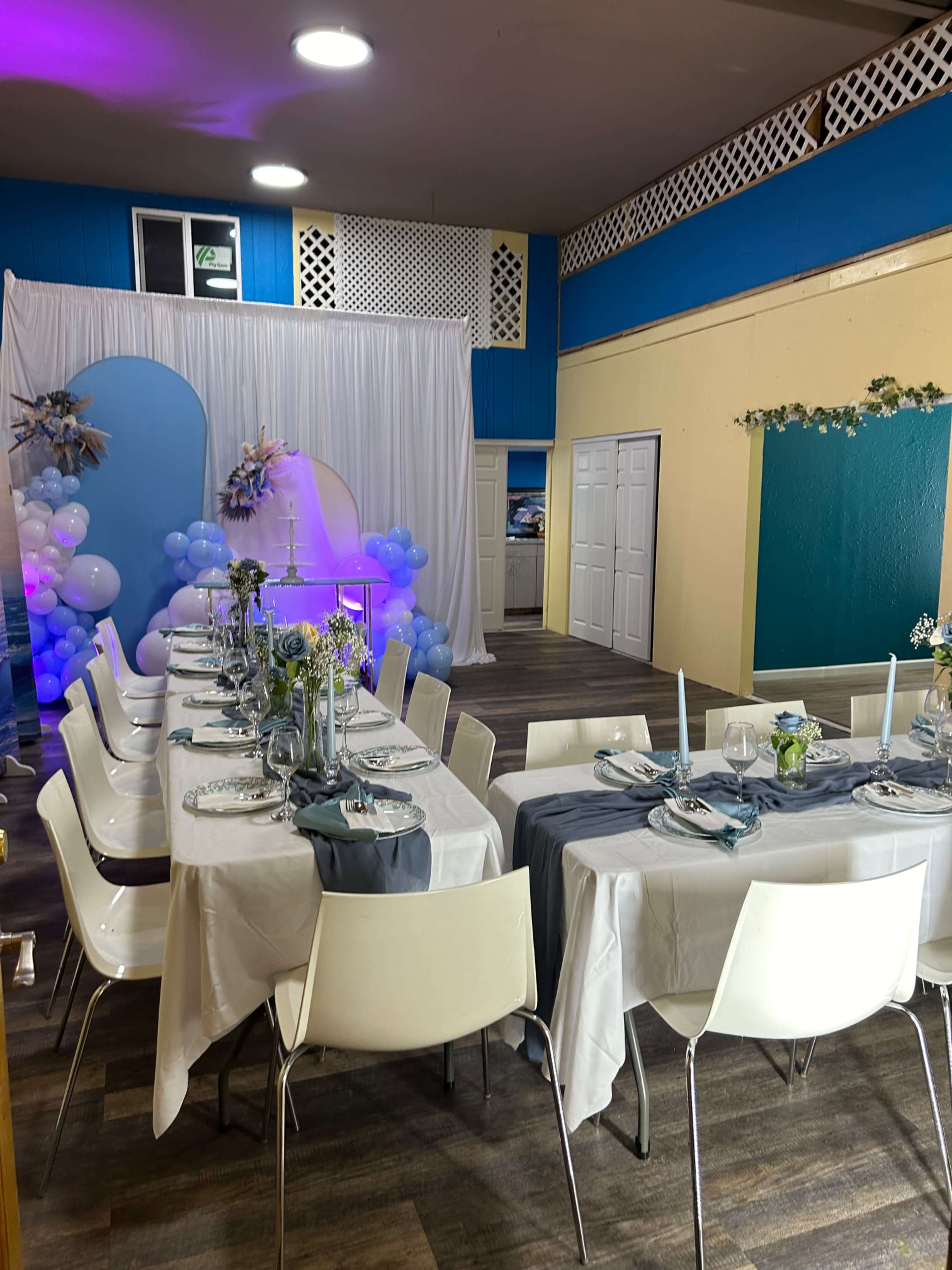 A decorated event space with two tables set for dining, featuring blue and white balloons, floral arrangements, and table settings with plates and glasses.