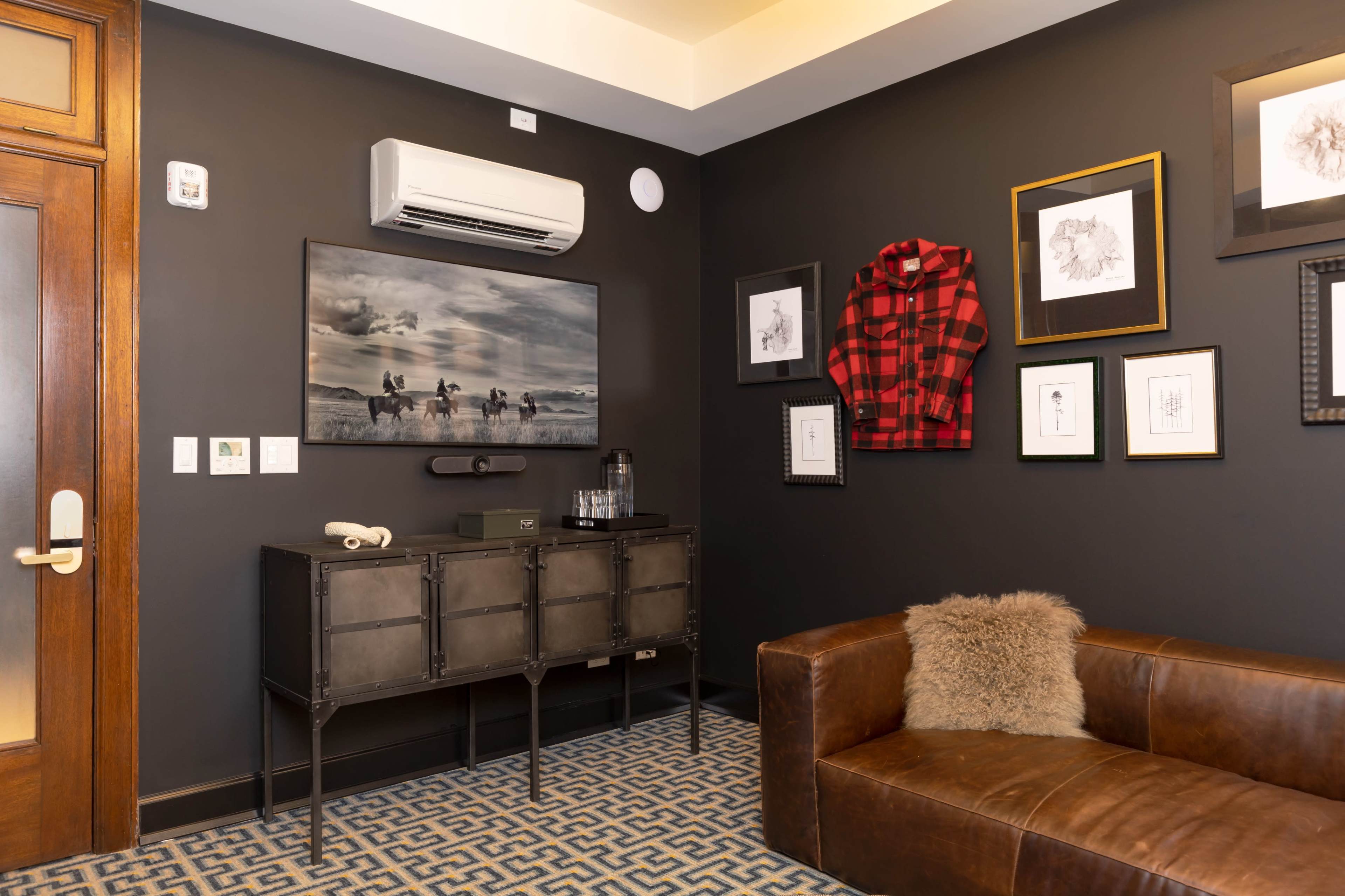 A cozy room with dark walls, a brown leather sofa, a patterned rug, framed artwork, and an air conditioning unit mounted on the wall.