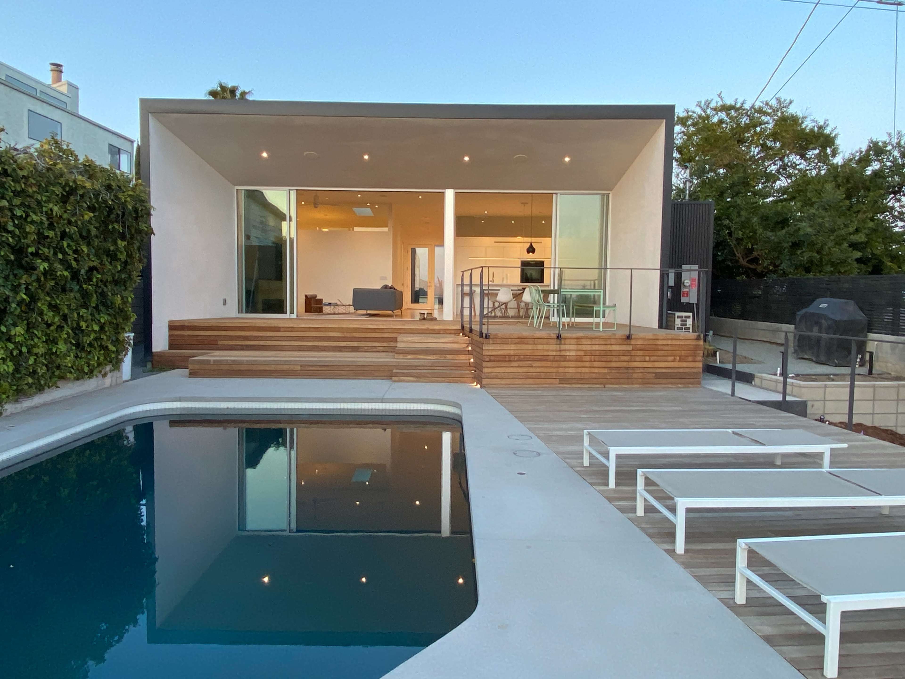 A modern house with large windows features a wooden deck and a swimming pool in the foreground.