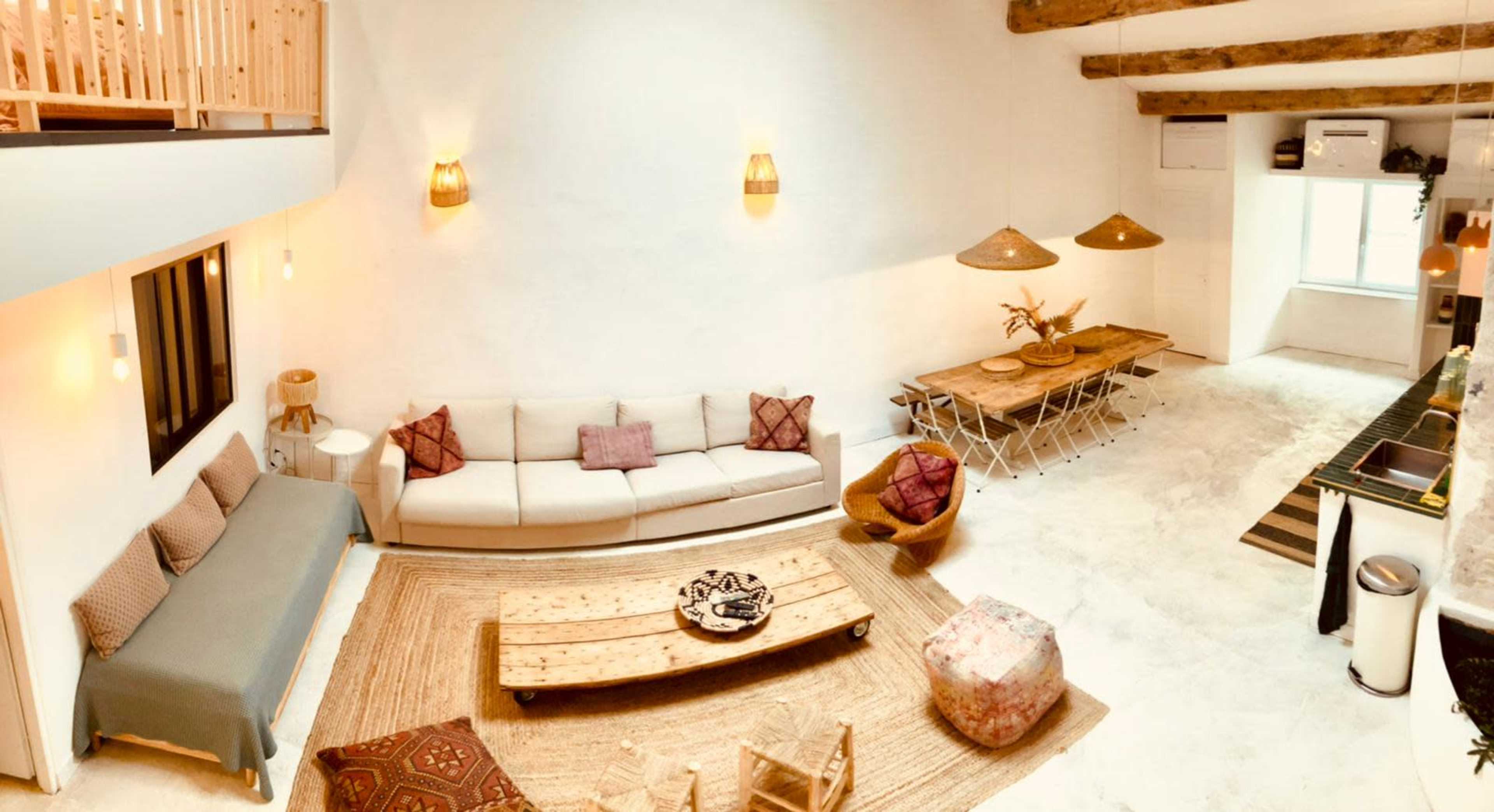 The image shows a contemporary, open-plan living area featuring a minimalist design with a sectional couch, a wooden coffee table, and a dining table with metal chairs.