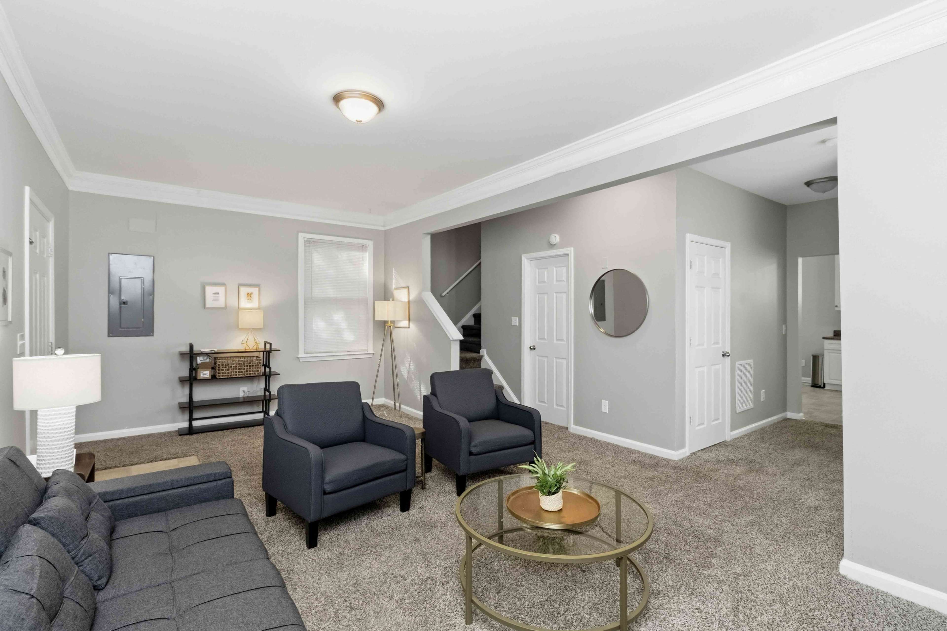 The living room features two dark chairs, a gray sofa, a round coffee table with a plant, and a view of a staircase and hallway.