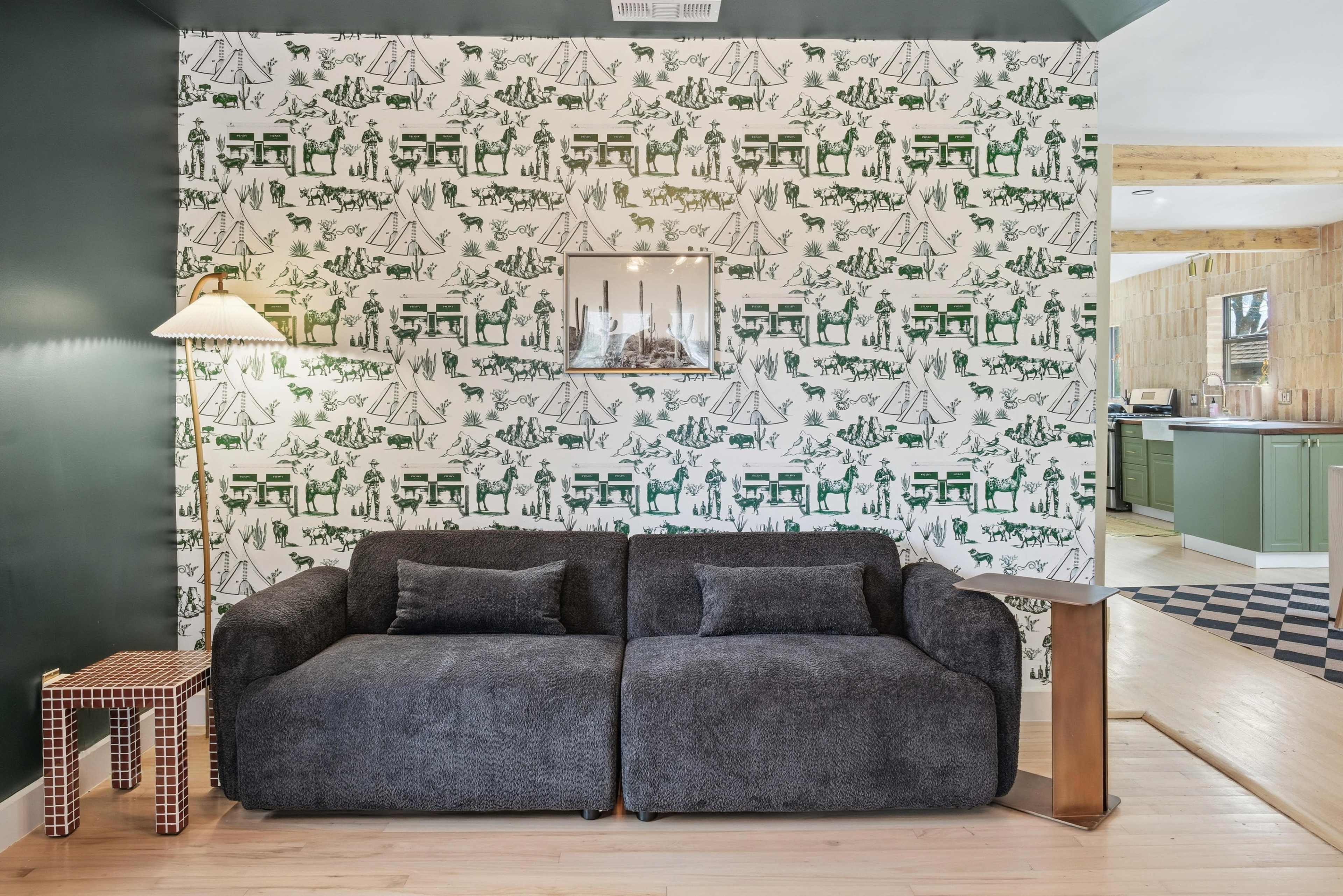 A dark gray sectional sofa faces a patterned green and white wallpaper, complemented by a lamp and a small checkered side table.