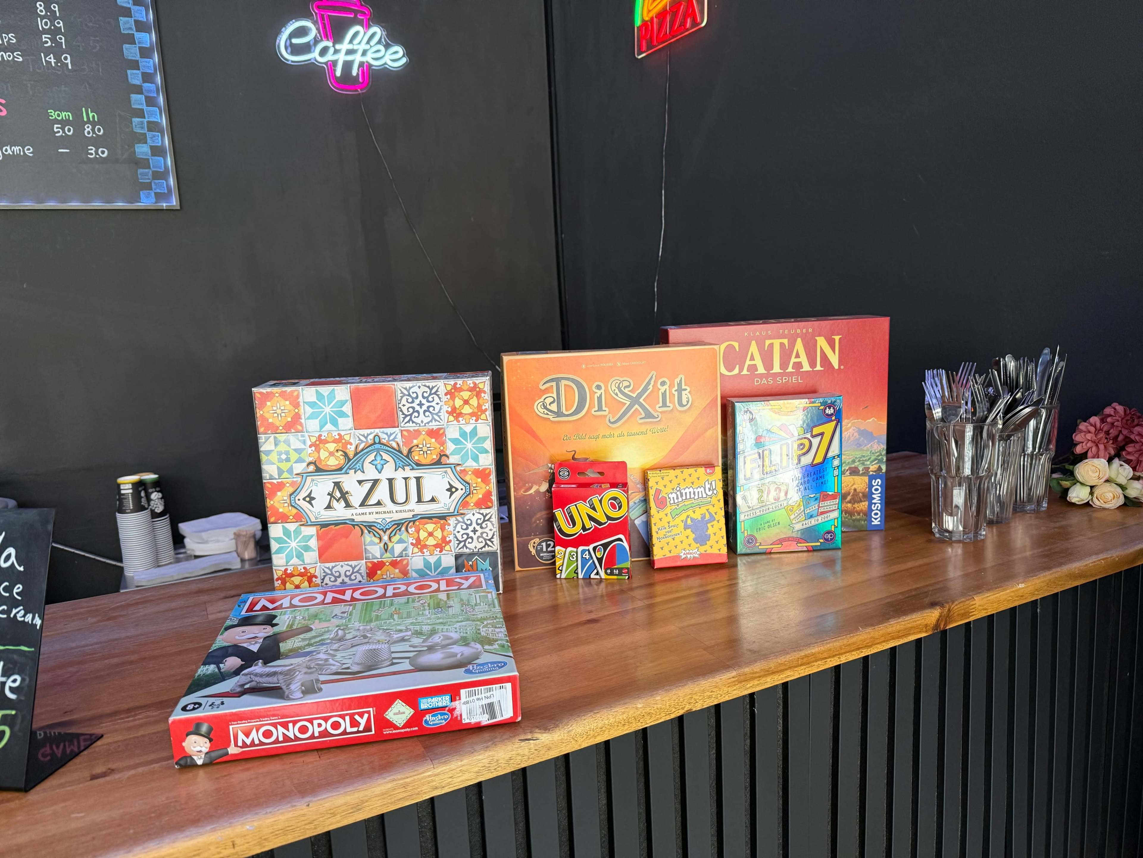 A wooden counter displays various board games, including Azul, Dixit, Catan, Uno, and Monopoly, alongside utensils in a casual setting.