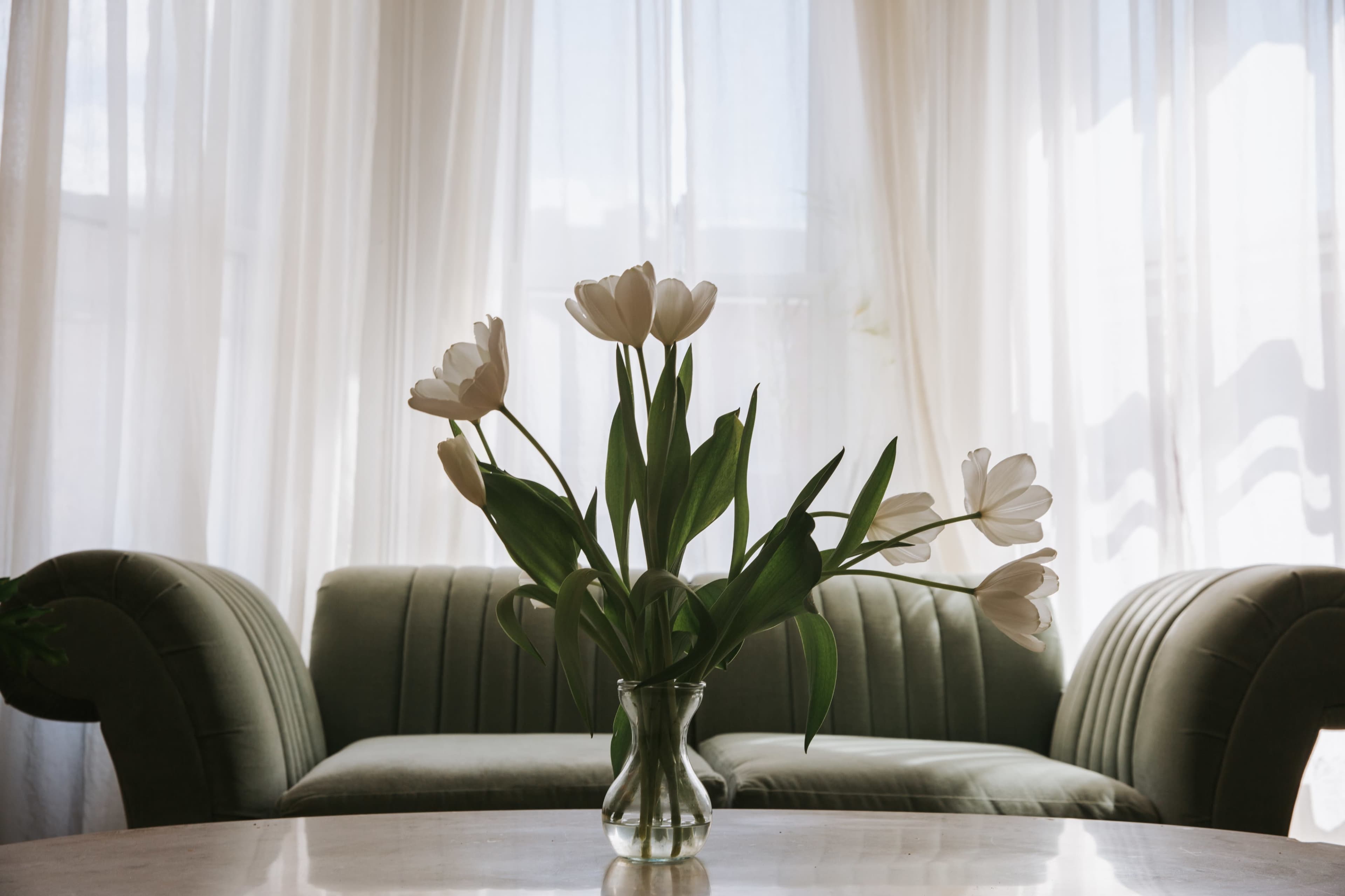 A vase of white tulips sits on a marble table in front of a green couch, with sheer curtains letting in natural light.
