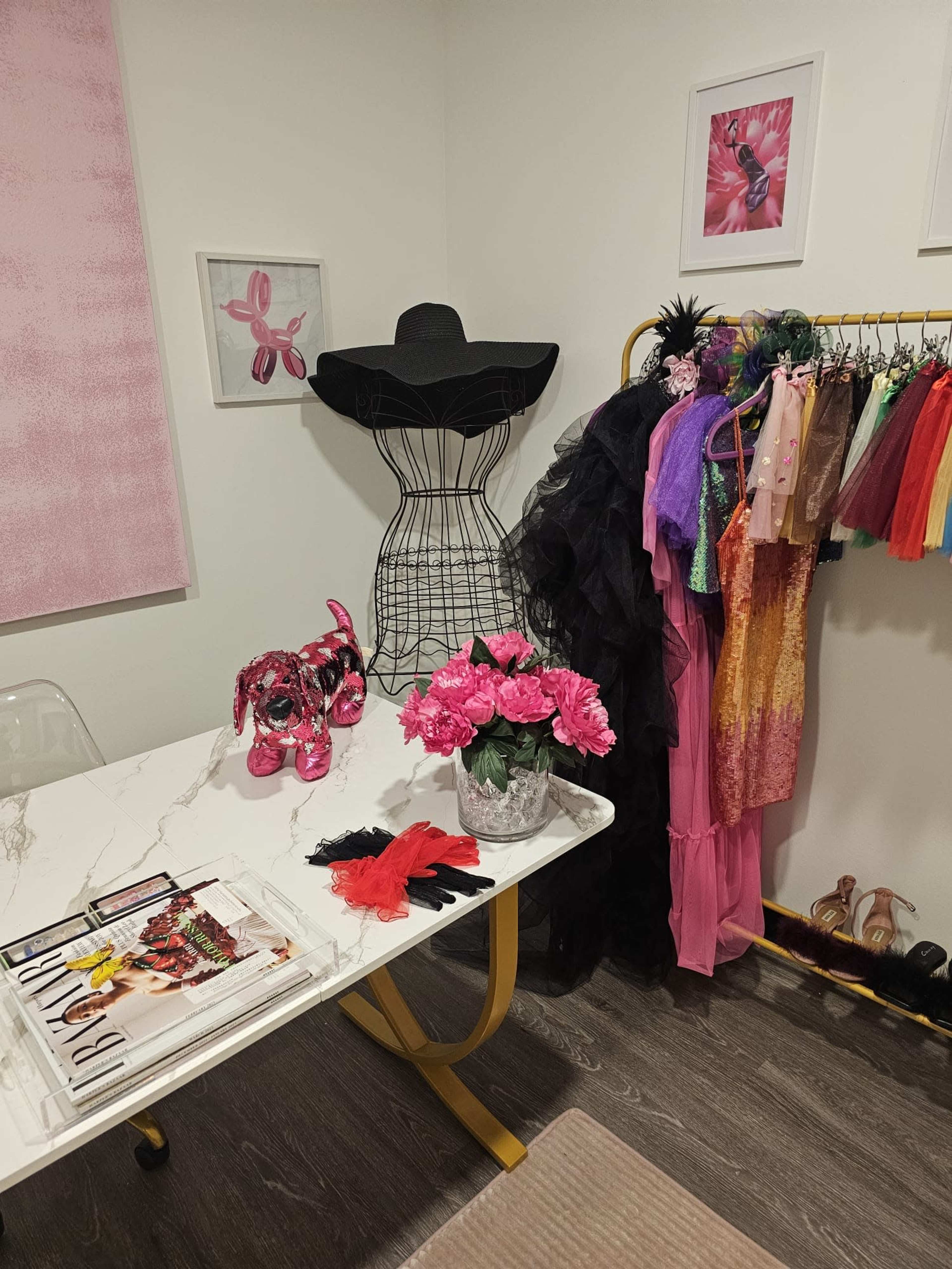A fashion styling workspace with a dress form, colorful clothing on a rack, a table with fashion magazines, and decorative flowers.
