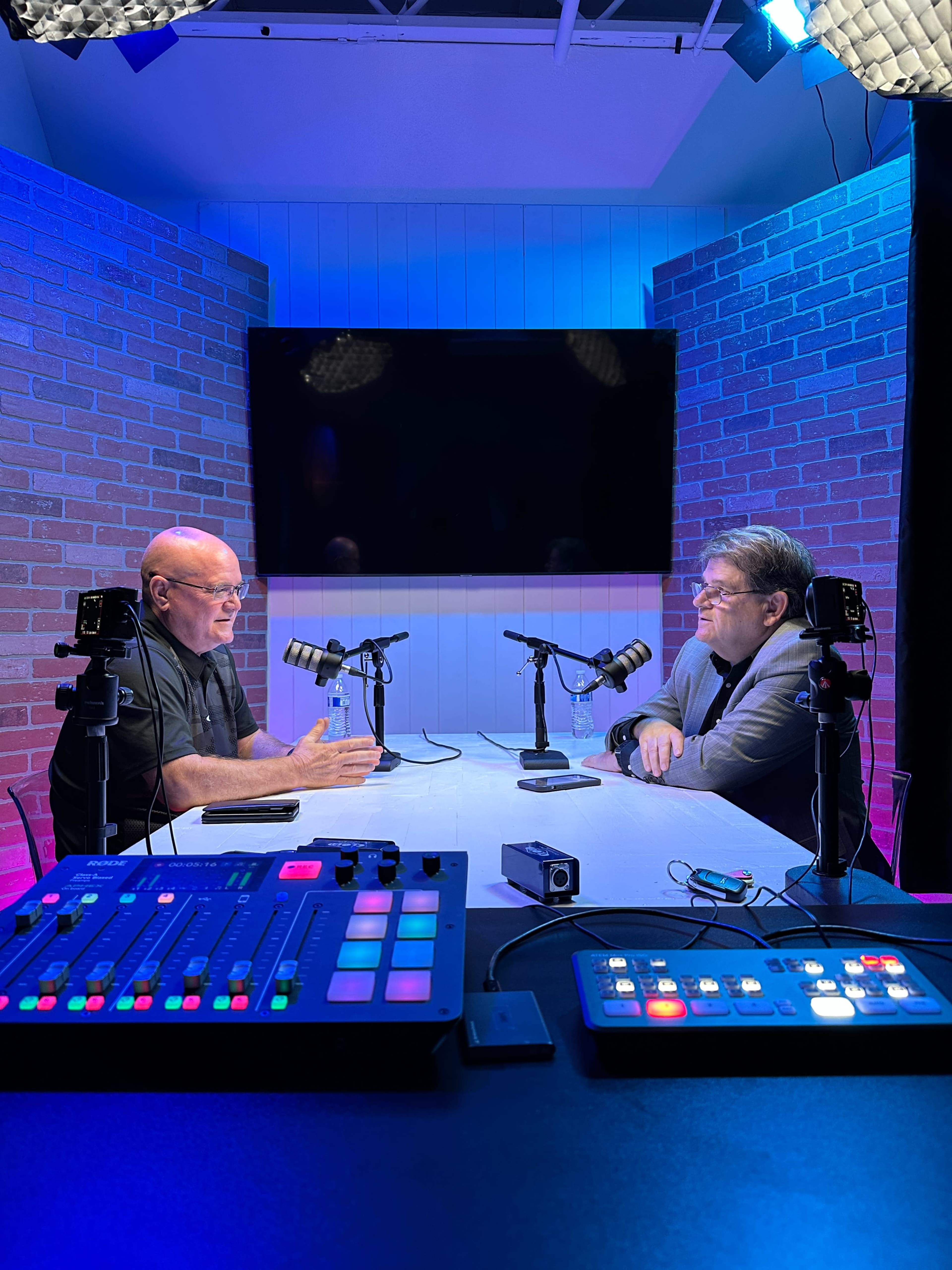 Two men are seated at a table with microphones, engaged in conversation, while a large screen and multiple lights illuminate the room.