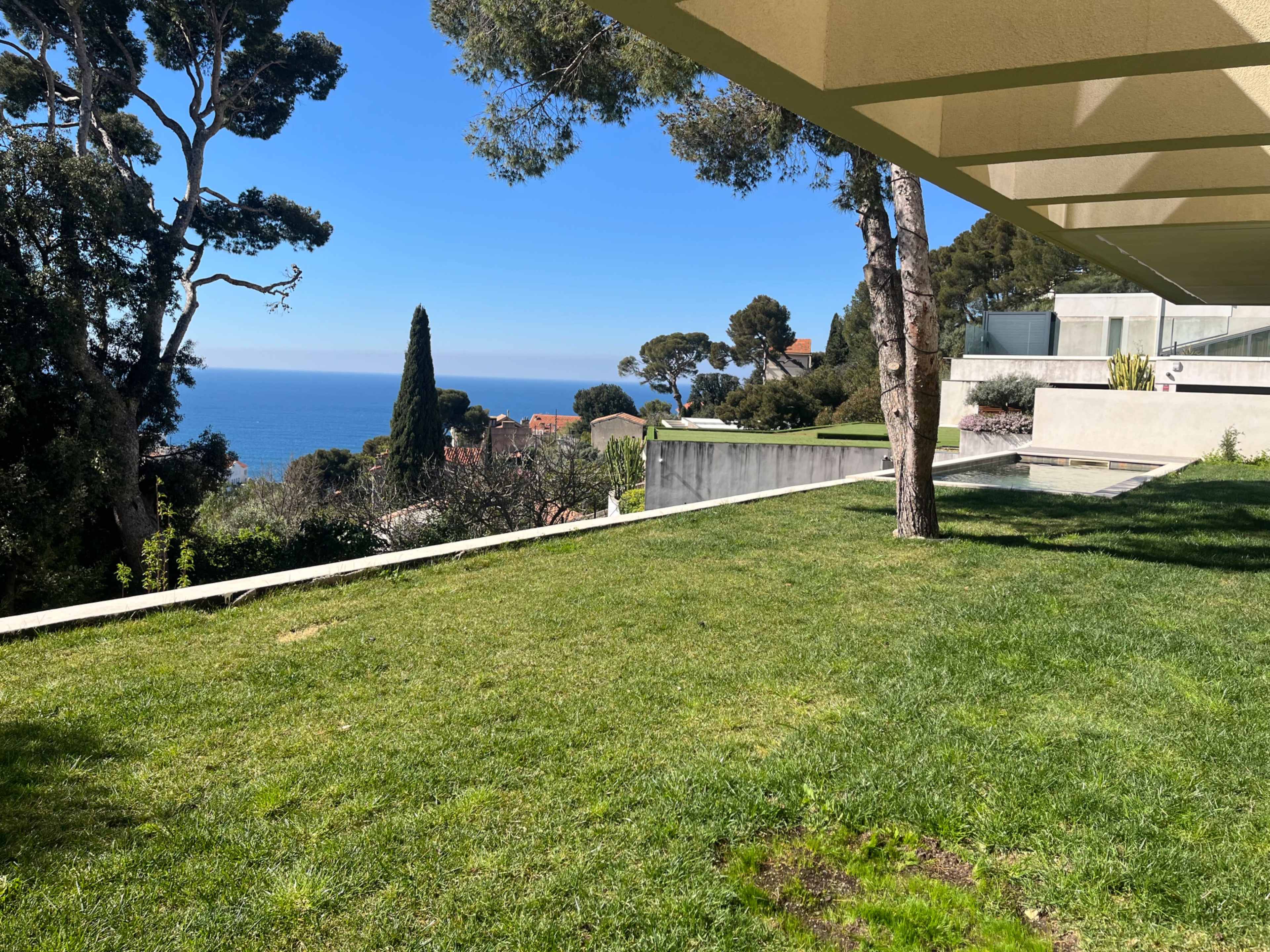 The image shows a grassy area with a clear view of the ocean and lush trees under a shaded architectural structure.