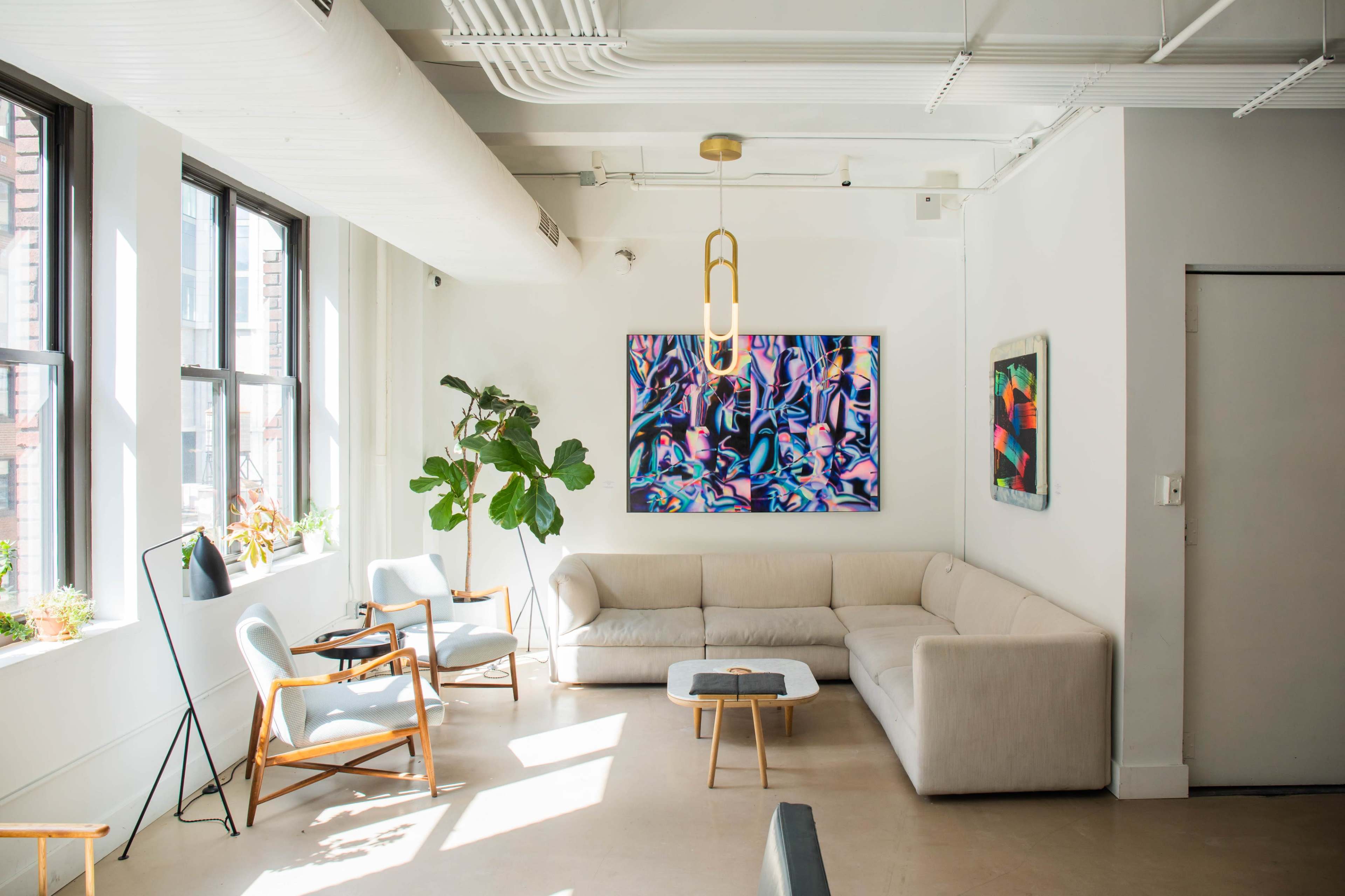 A bright, modern living space with large windows, a beige sectional sofa, and two light blue chairs, featuring colorful artwork on the walls.