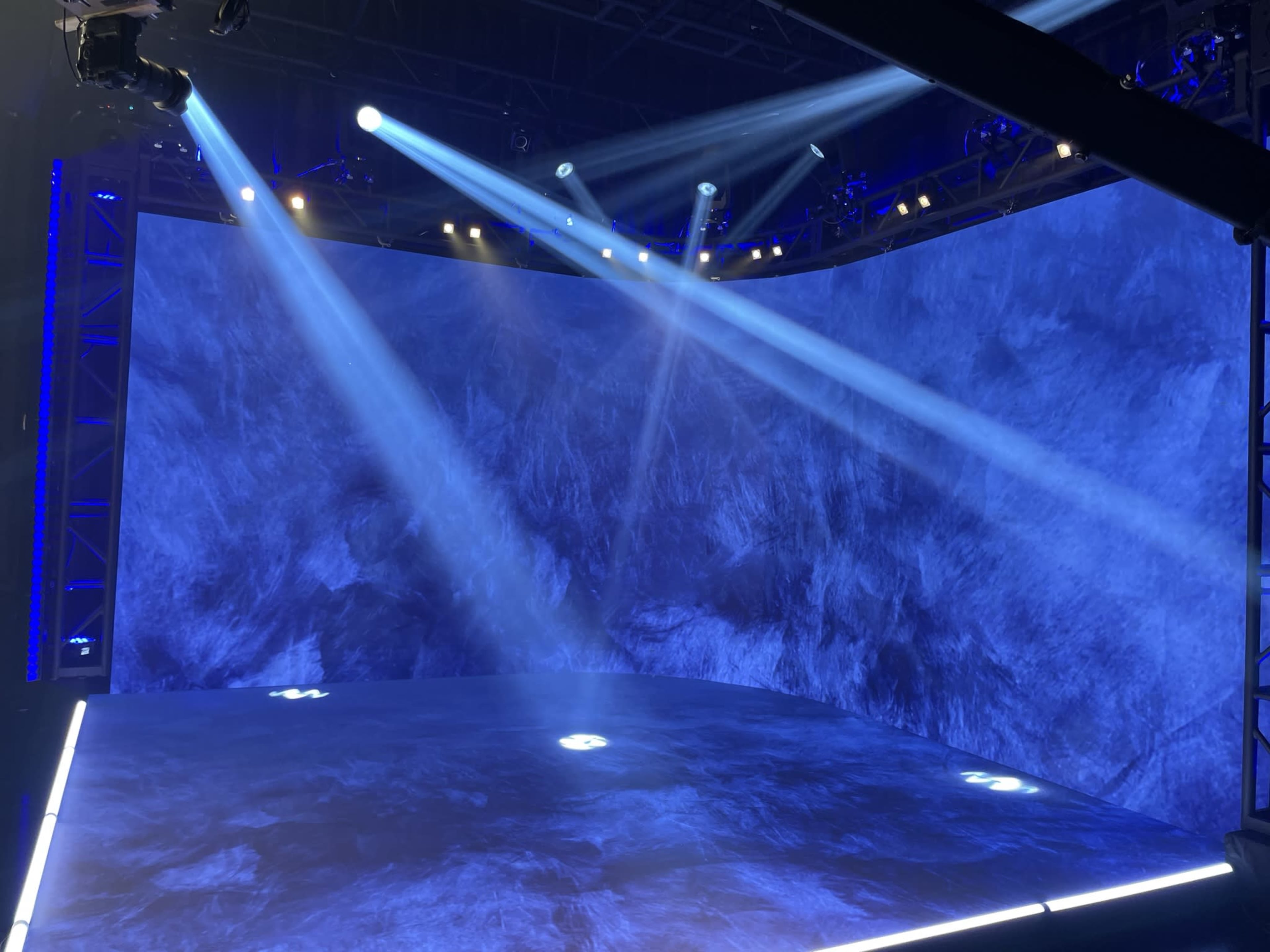 A stage with a curved blue backdrop, illuminated by beams of light from overhead fixtures.