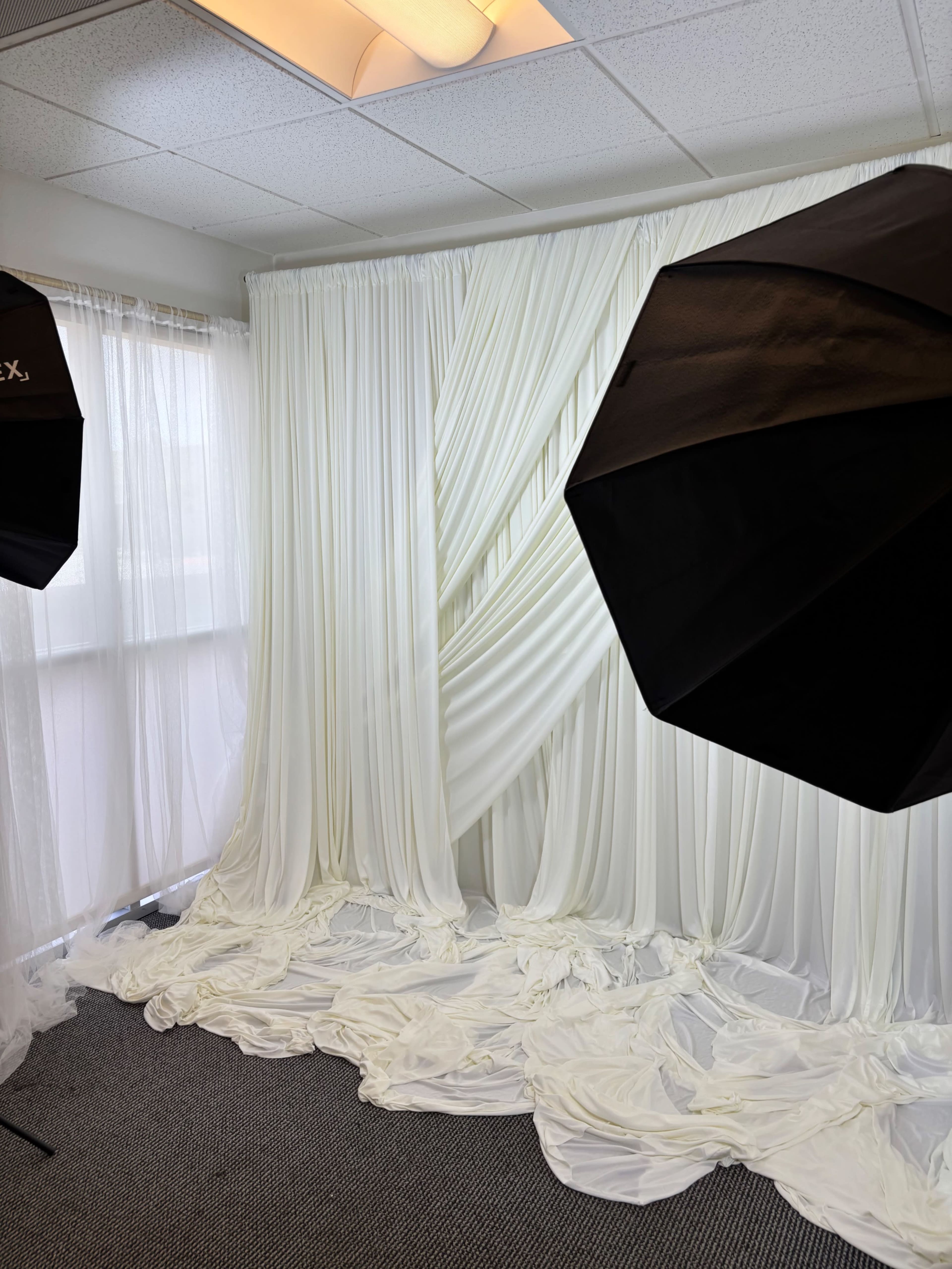The image shows a photography studio setup with white draped fabric as a backdrop and two softbox lights positioned to illuminate the space.
