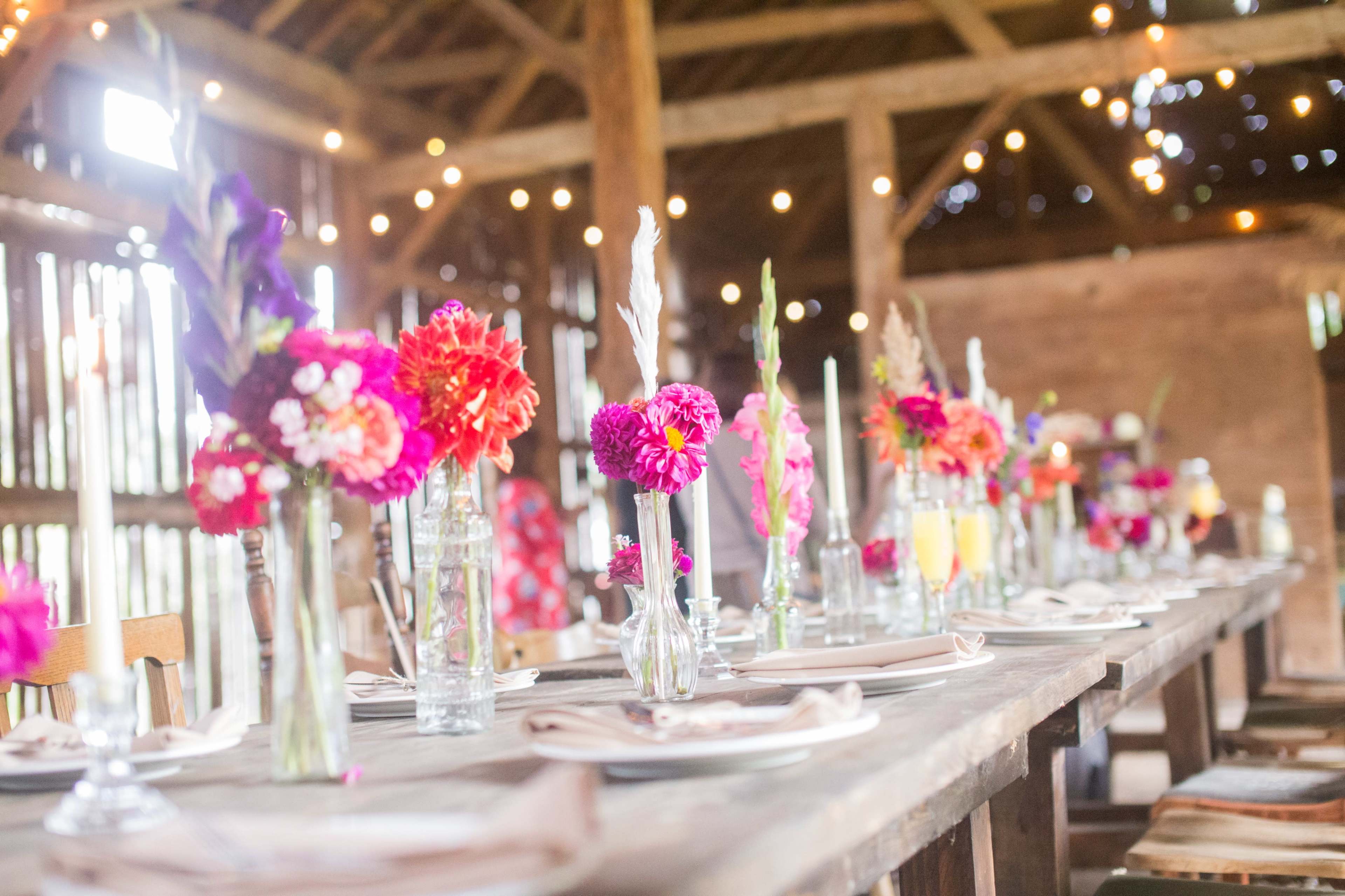 A long wooden table is set with colorful floral centerpieces and glass bottles, illuminated by string lights inside a rustic barn.