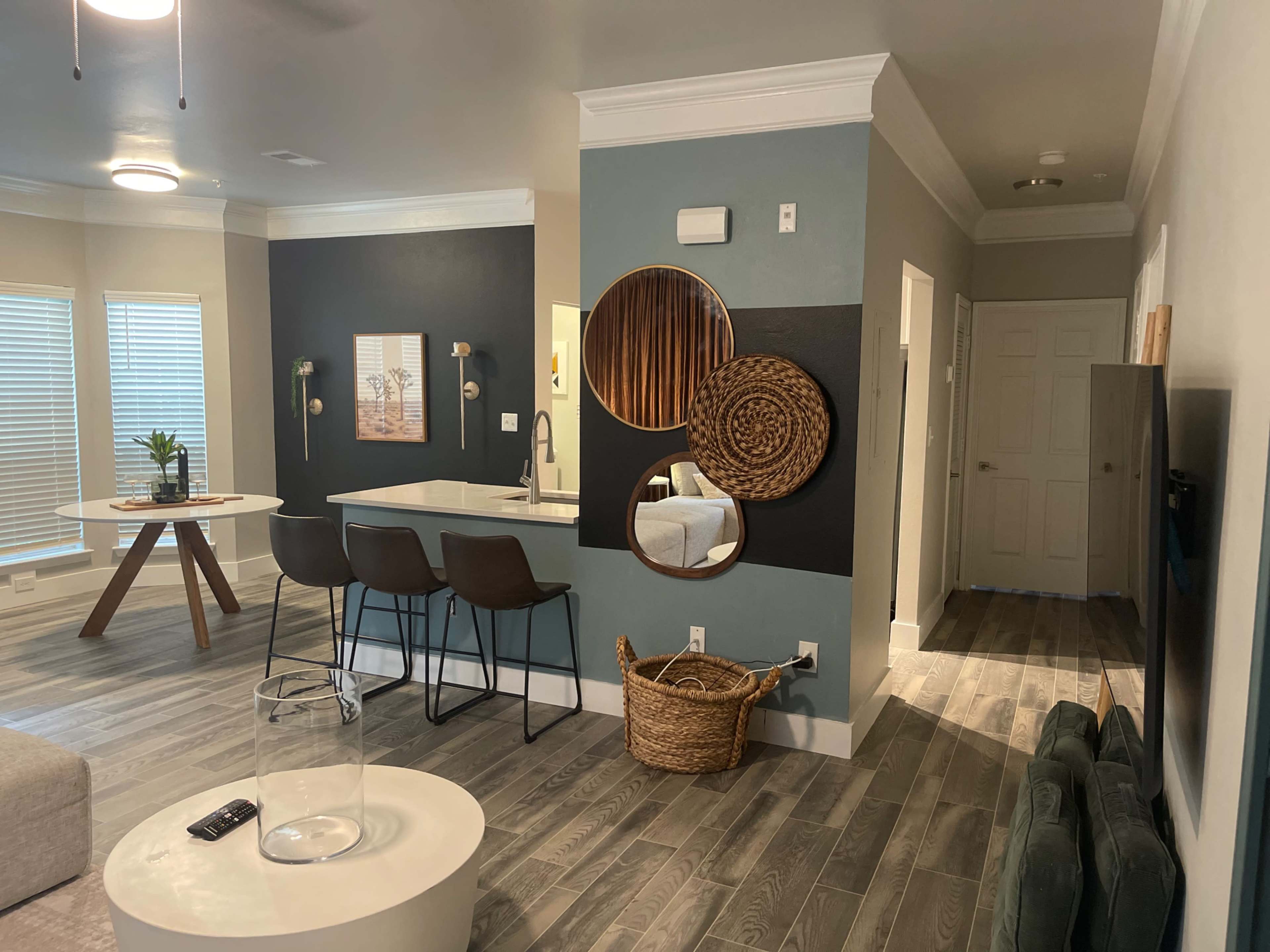A modern living area with a circular dining table, bar stools at a kitchen counter, and a wall adorned with decorative mirrors and woven baskets.