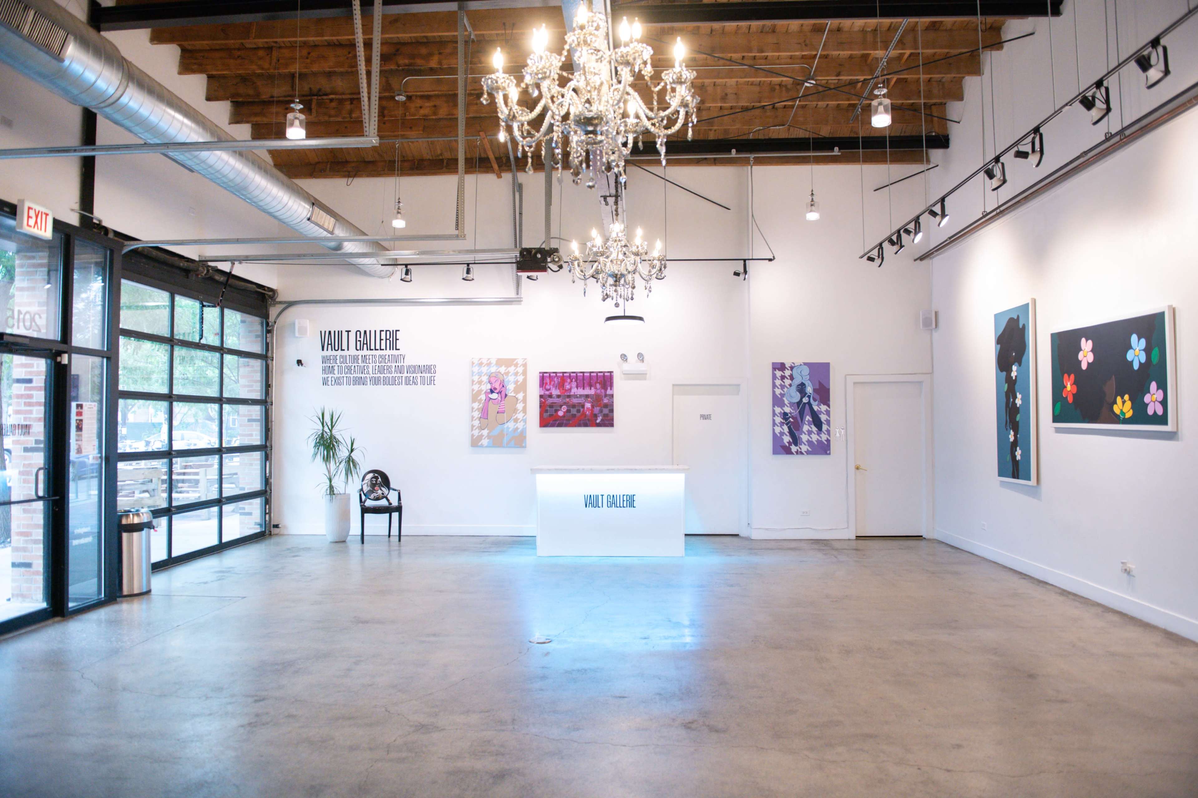 The image shows a spacious art gallery featuring white walls, modern artworks, a chandelier, and a central illuminated display area.