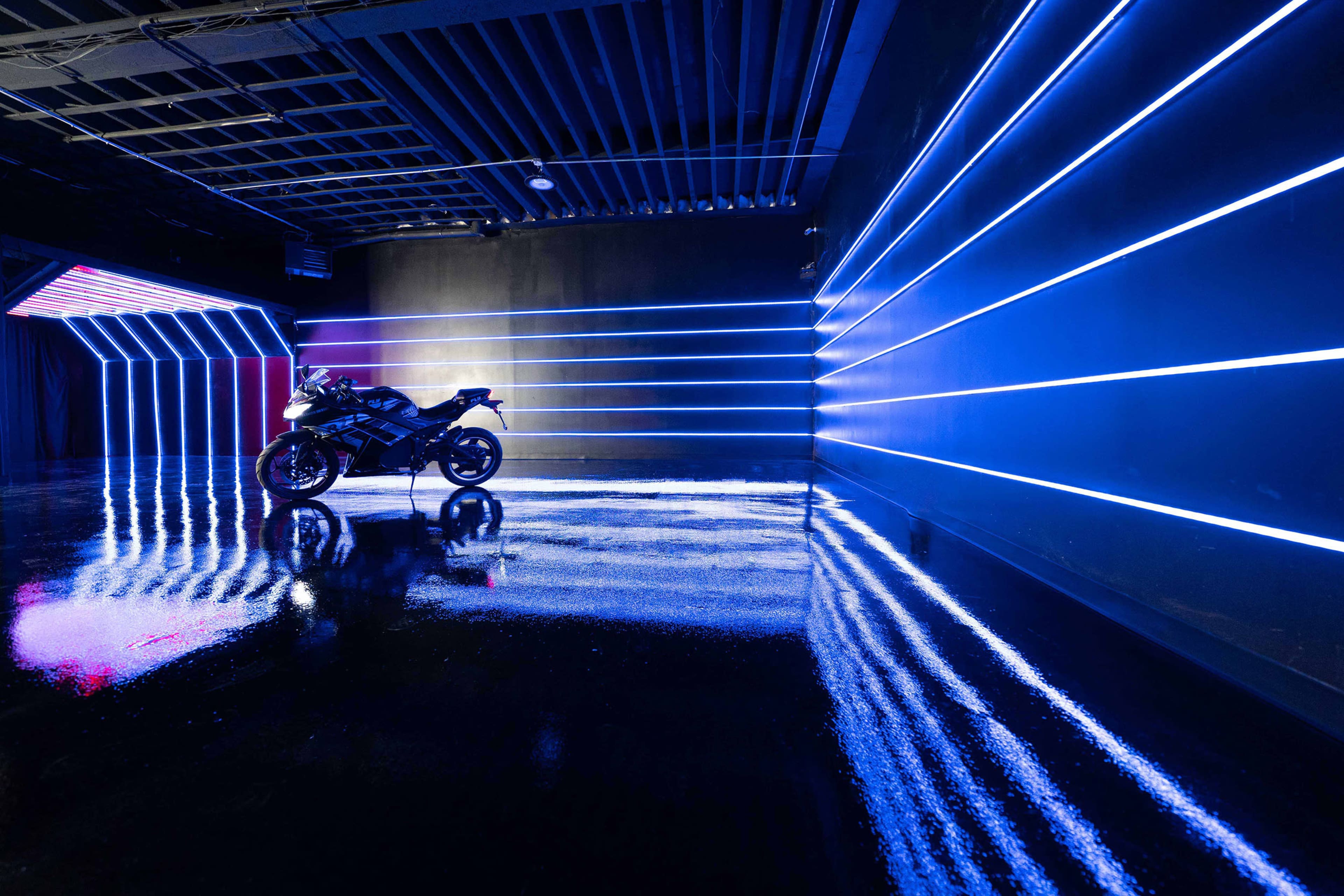 A motorcycle is positioned in a dark room illuminated by blue neon lights creating geometric patterns on the walls and floor.