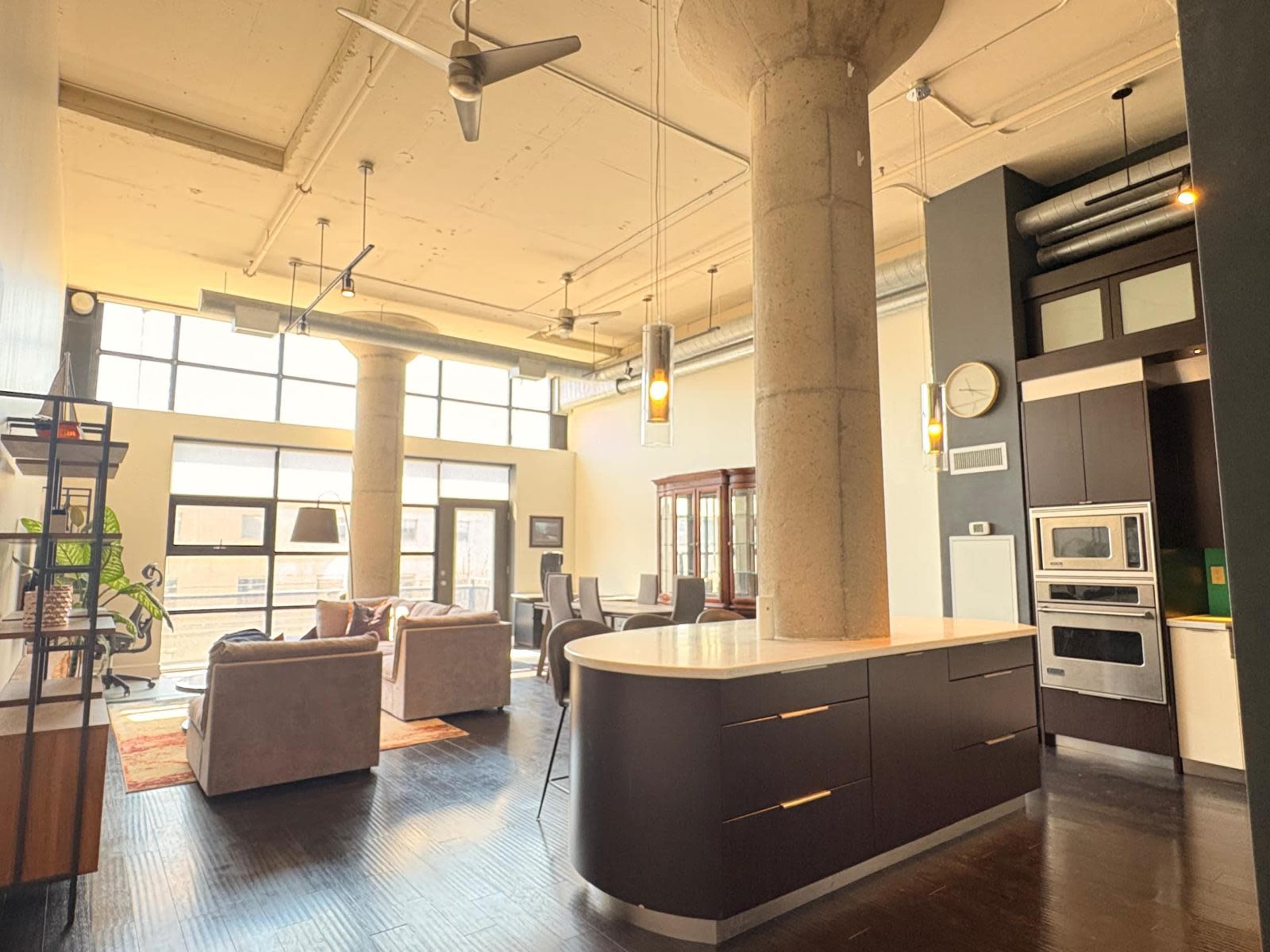 The image shows a modern open-concept living space featuring a kitchen with dark cabinetry, a dining area, and a seating area with large windows that allow natural light to fill the room.