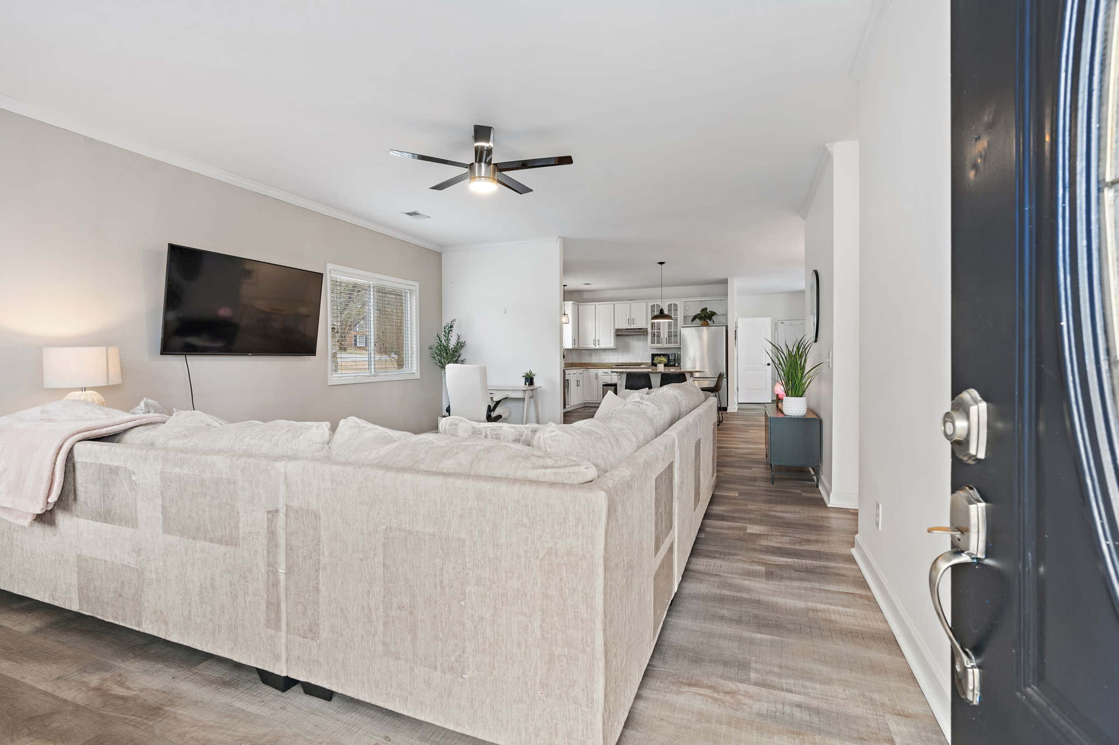 The image shows a living room with a large sectional couch, a wall-mounted television, and an open view into a kitchen area with modern appliances.