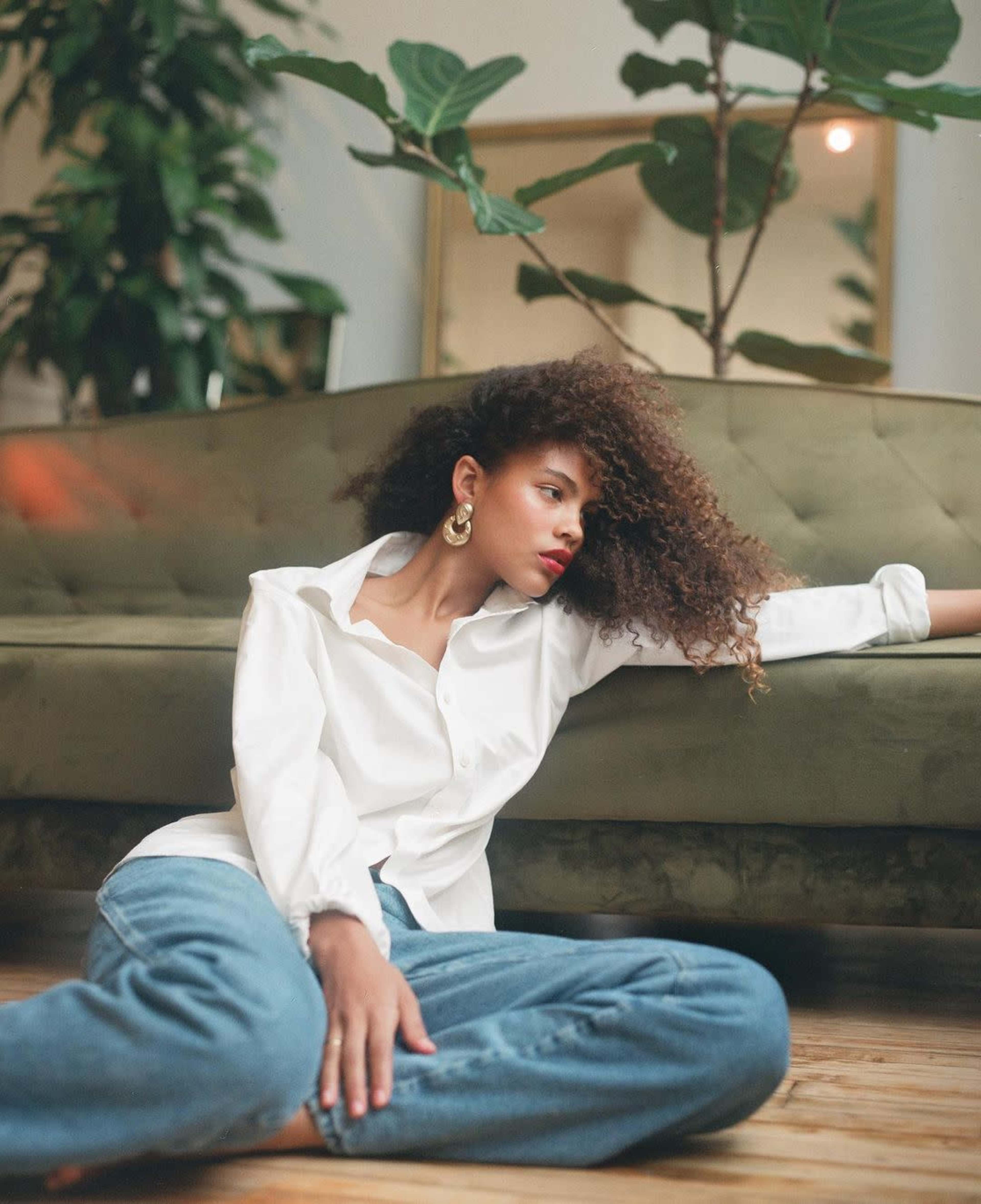A person with curly hair is seated on a green sofa, wearing a white button-up shirt and blue jeans, with a large plant in the background.