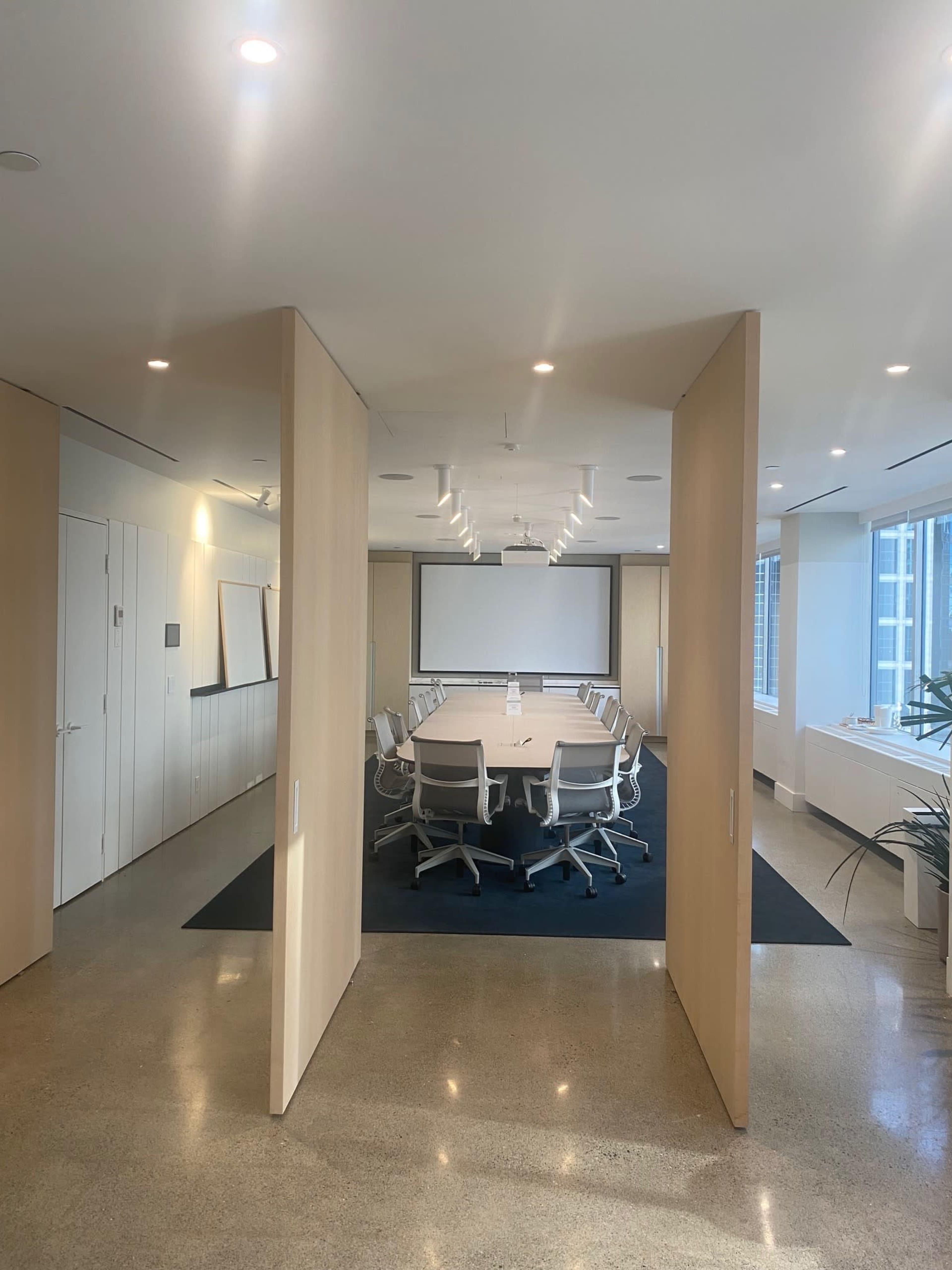 A modern conference room with a large table surrounded by white chairs, partitioned by wooden walls, and equipped with presentation technology.