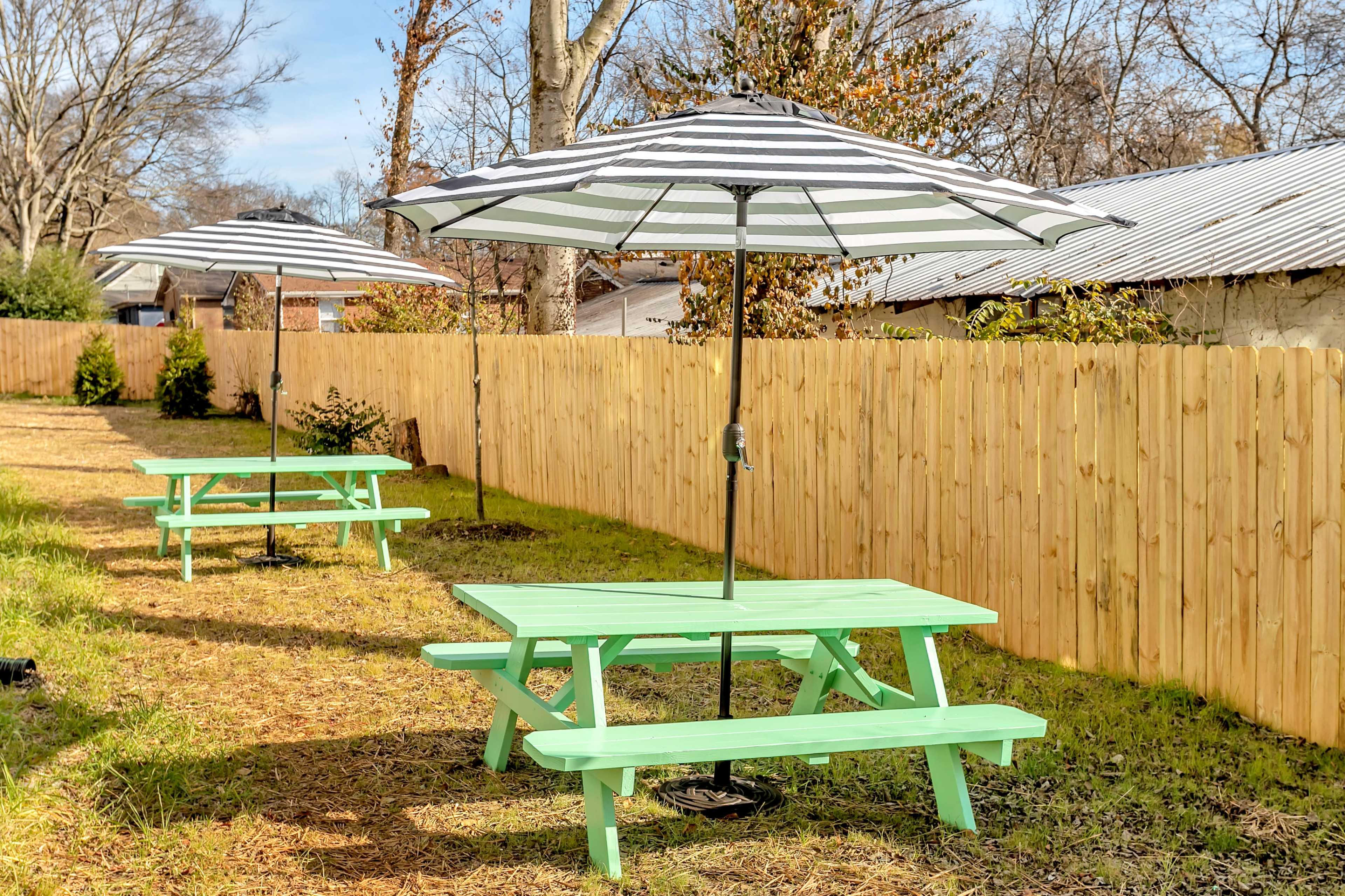Two green picnic tables with striped umbrellas set in a grassy area next to a wooden fence.