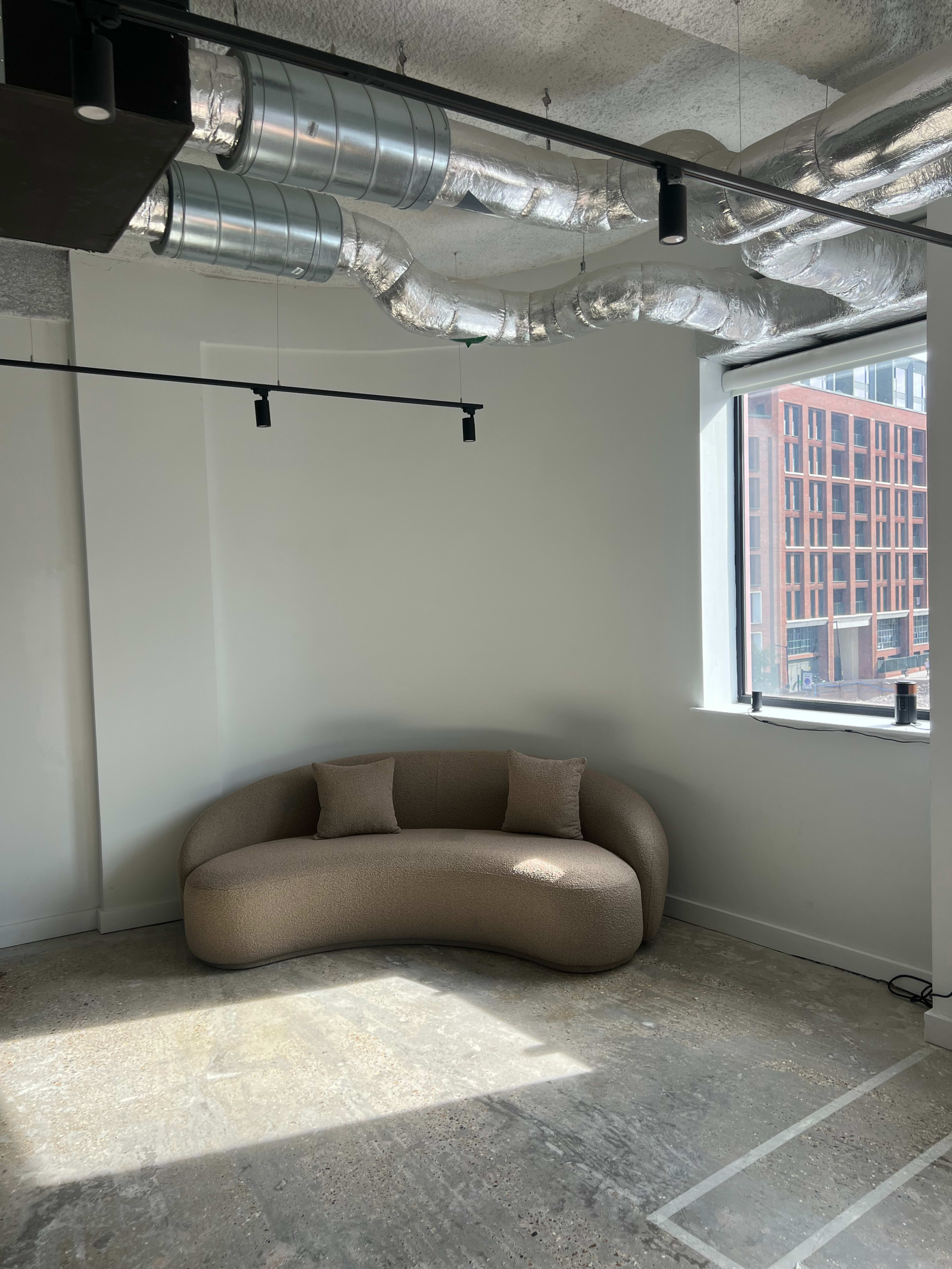 A curved, beige sofa with two throw pillows is positioned against a wall in a sunlit, minimalist office space.