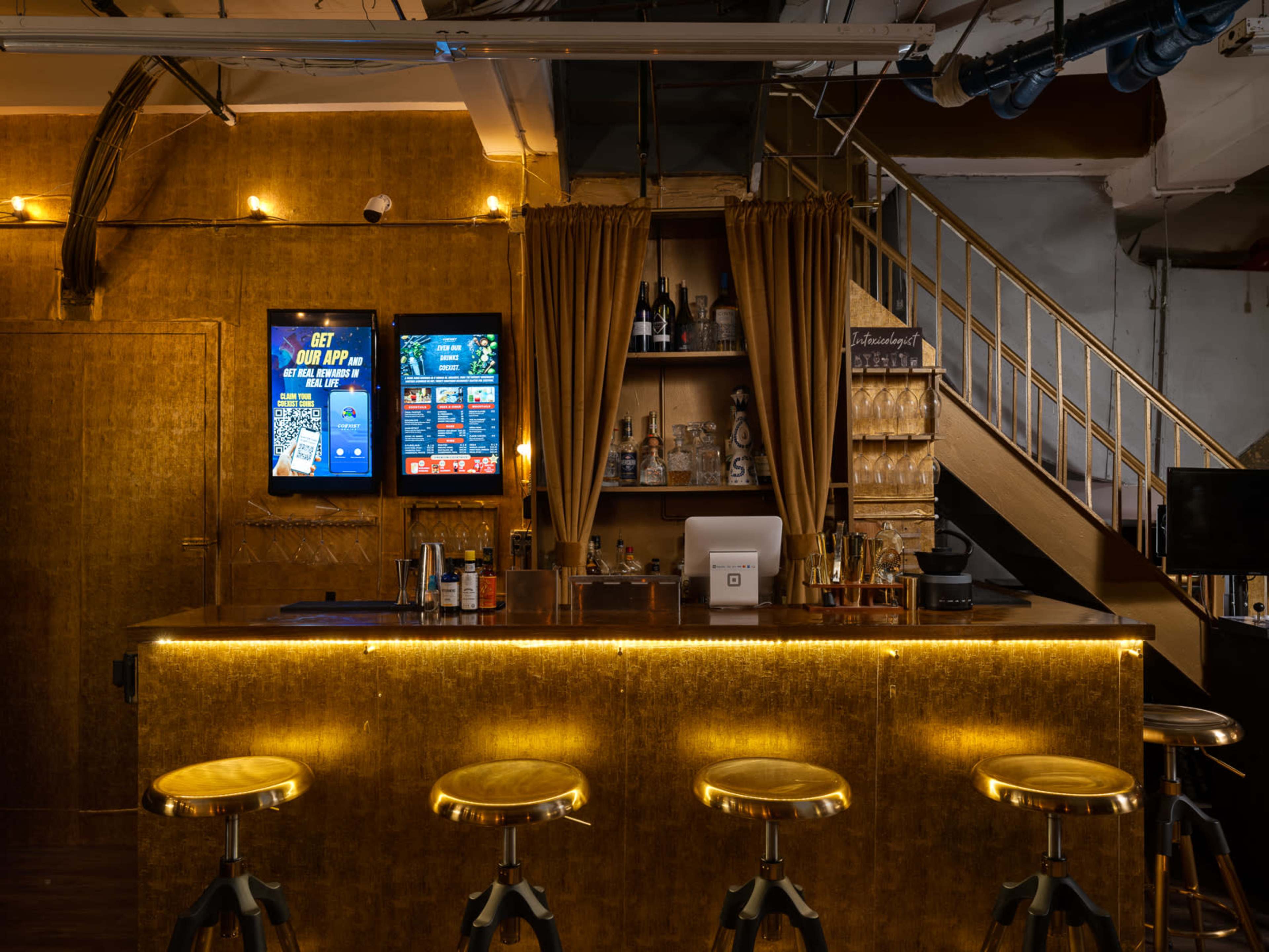 The image shows a bar with a golden-hued counter, four high stools, a digital display, and a staircase in the background.