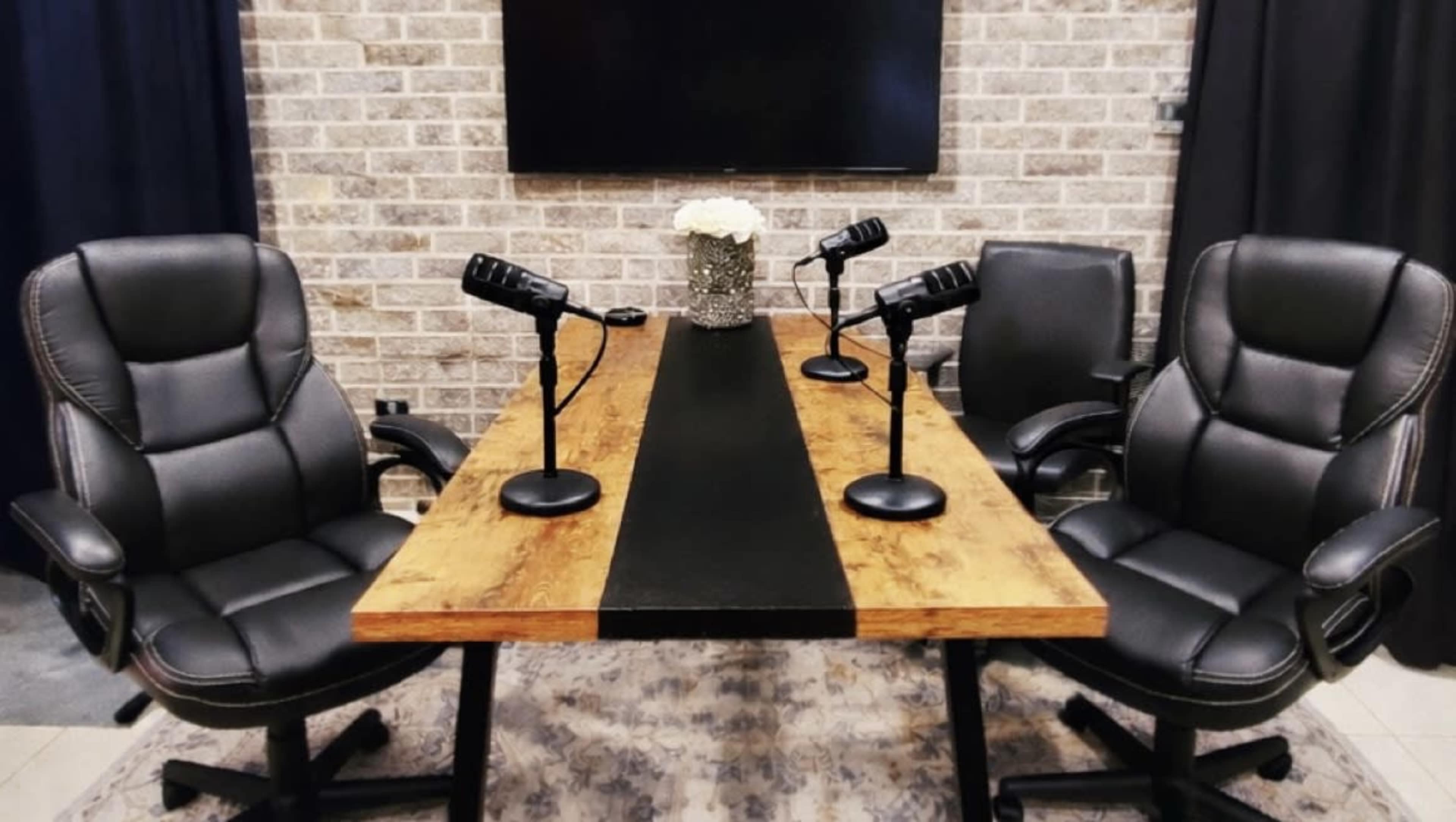 The image shows a podcast recording setup with two black leather chairs at a wooden table, each equipped with a microphone, against a brick wall background.
