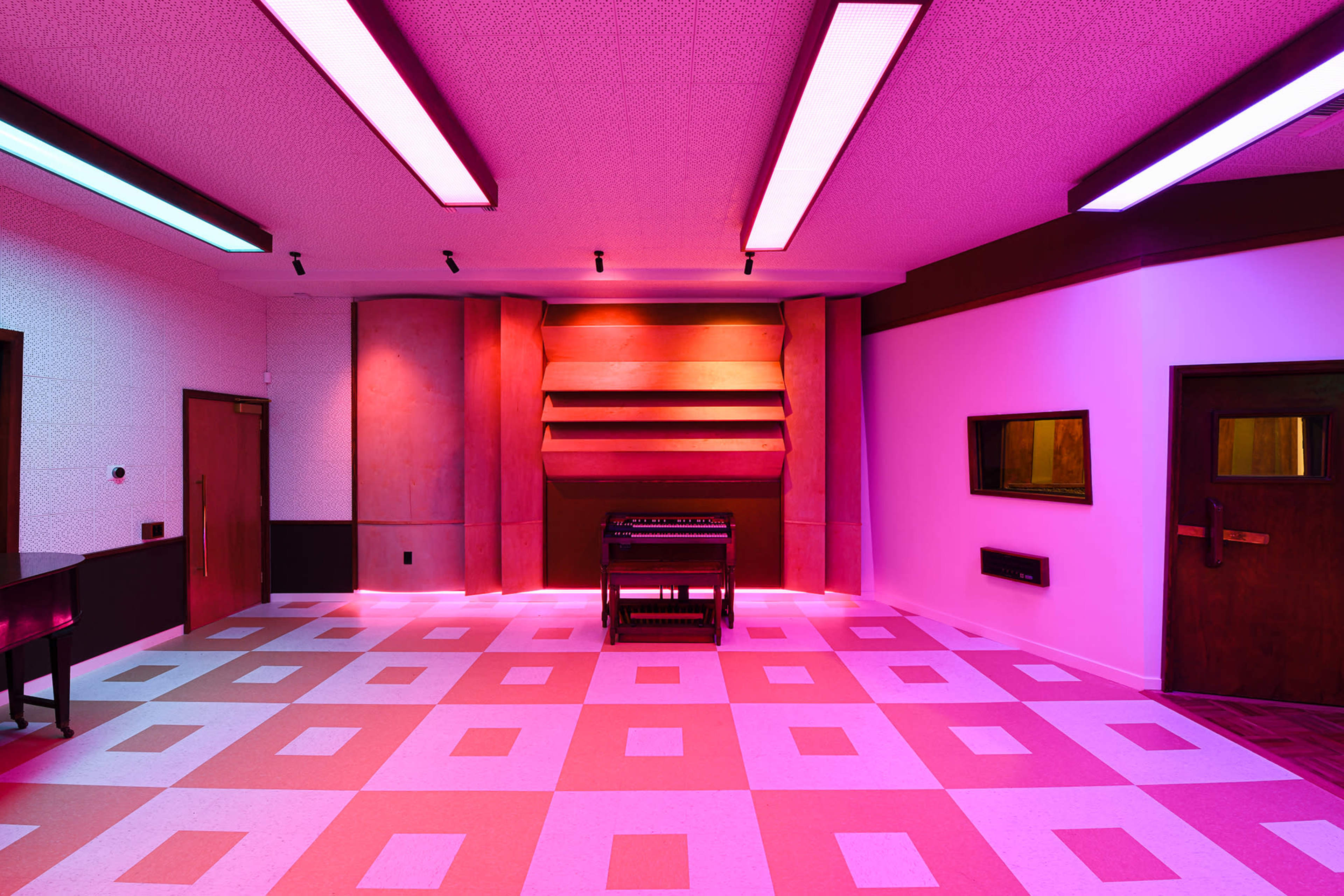 A brightly lit room with pink and purple lighting, featuring a patterned floor and an organ in the center against a stylized wall.