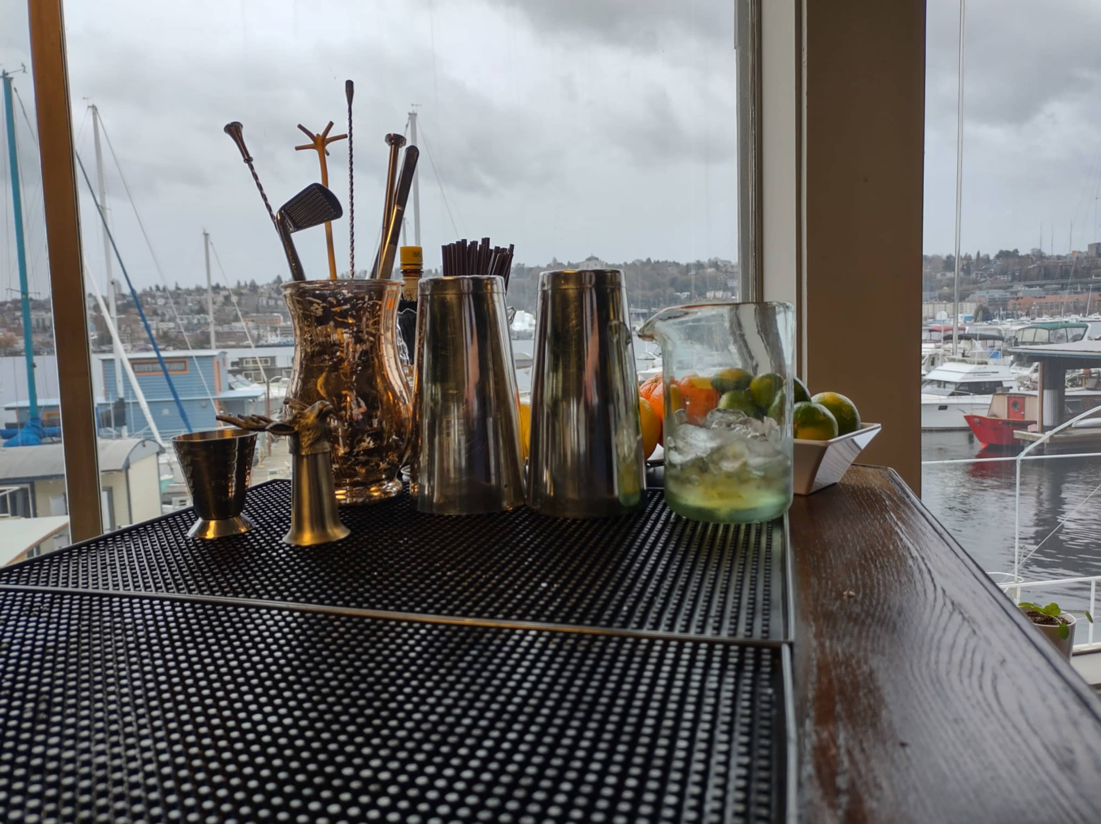 A bar counter displays various cocktail tools and a pitcher next to a glass of drink, with boats visible outside the window.