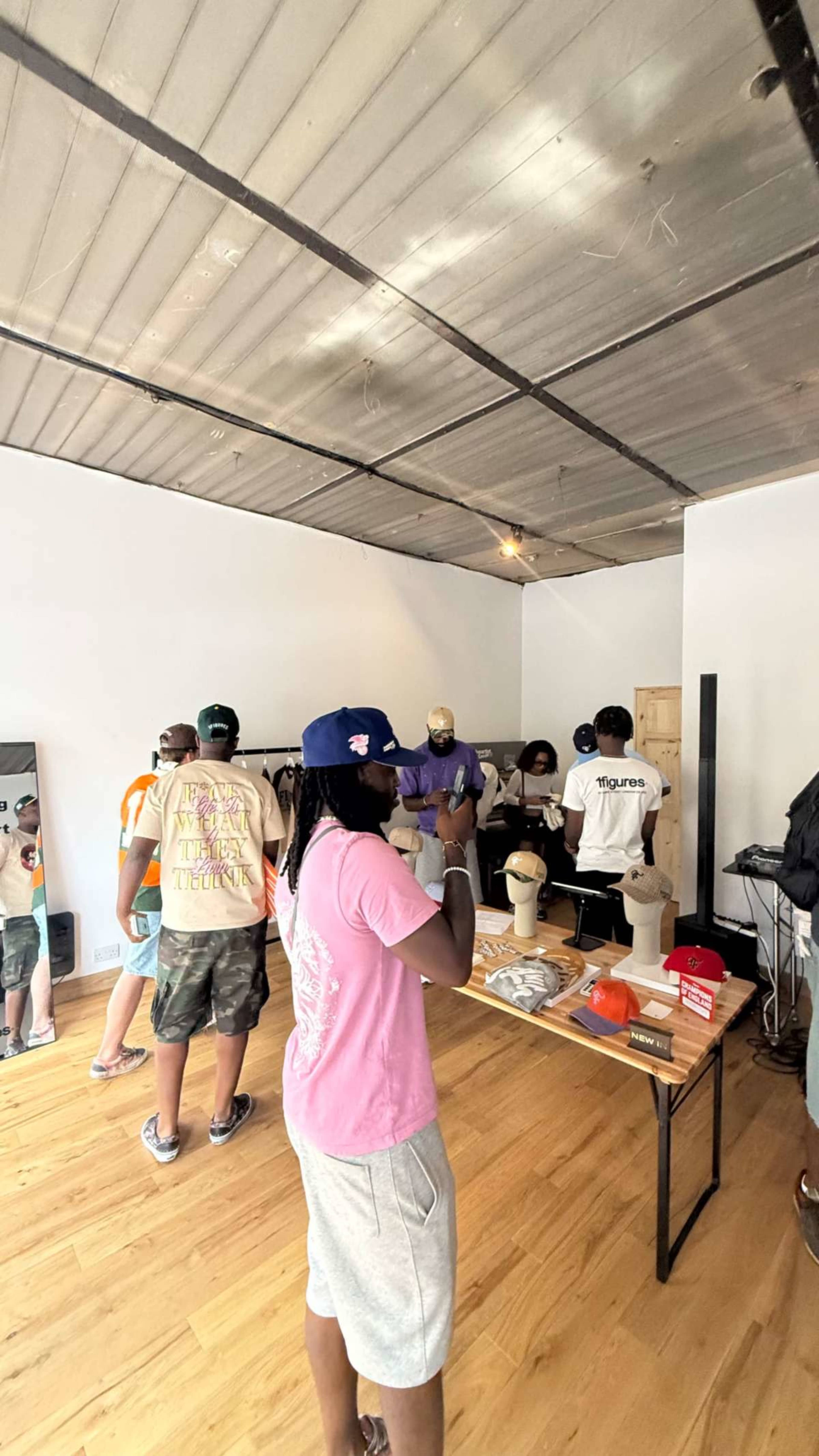 A group of people is standing in a bright, spacious room with wooden flooring, engaging with a display table and trying on hats and accessories.