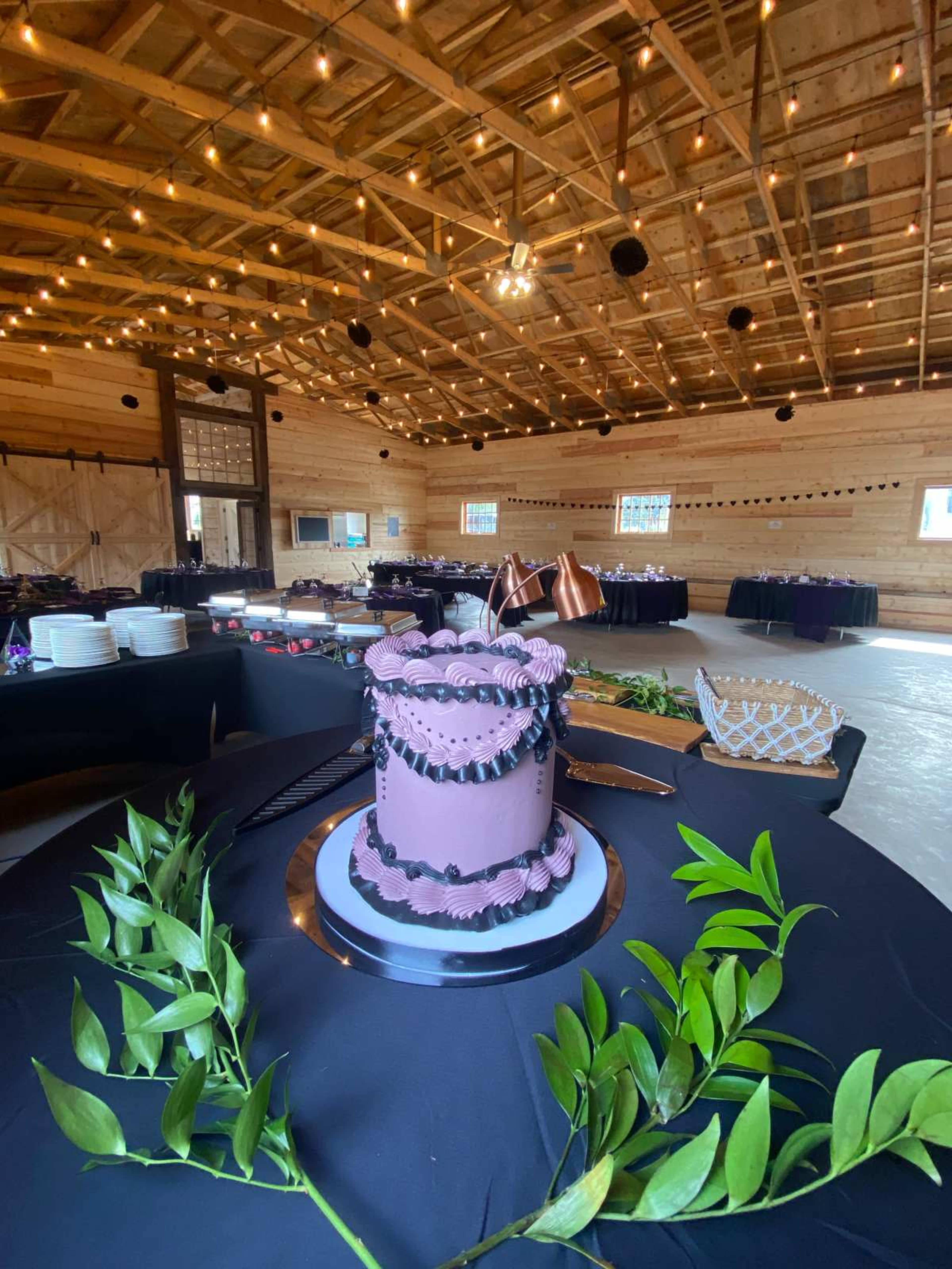 The image shows a decorated cake at the center of a dining setup inside a wooden barn, with string lights illuminating the space and tables covered in black tablecloths.