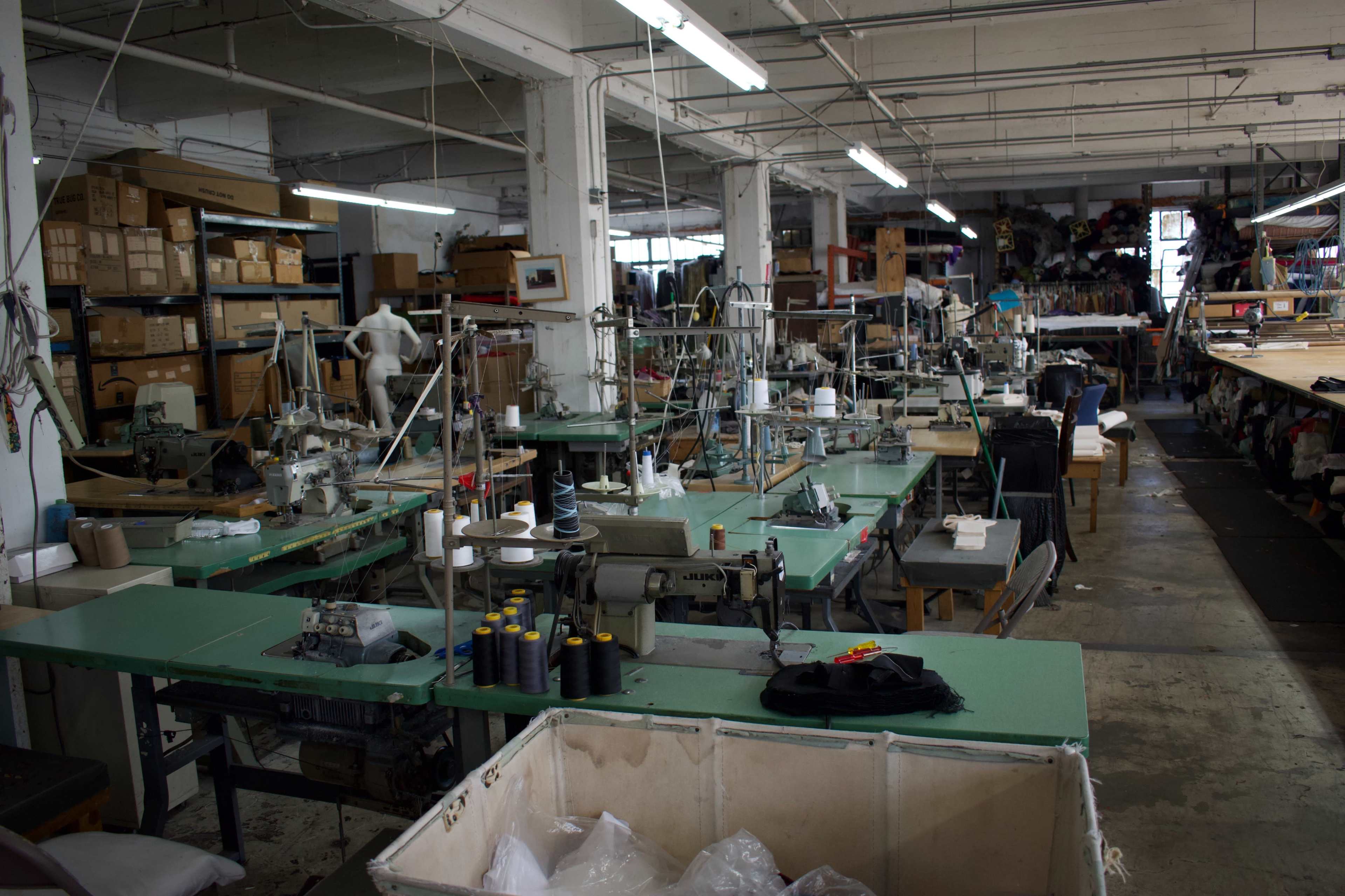 The image shows a spacious sewing workshop filled with sewing machines, work tables, and stacks of boxes.