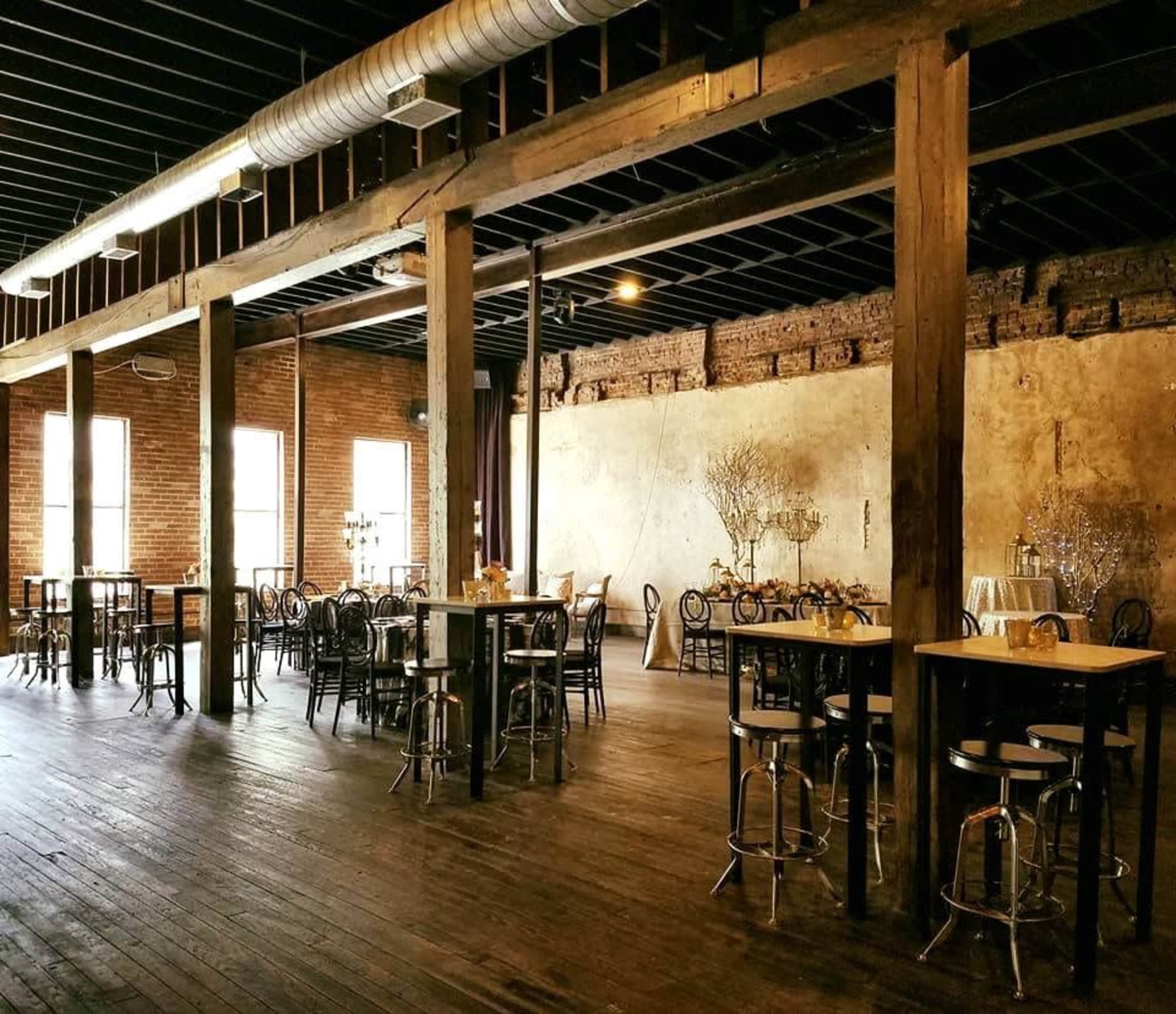 An empty industrial-style event space with wooden beams, brick walls, and several tables scattered throughout.