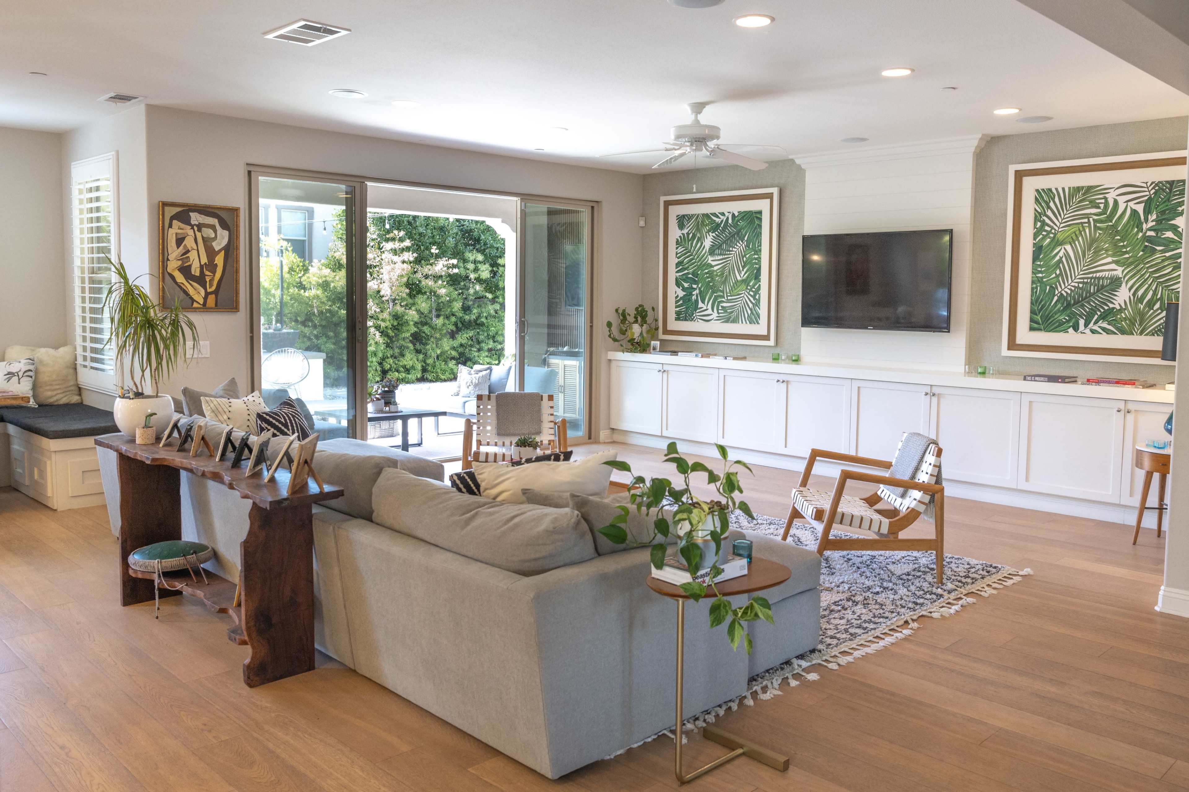 A modern living room with a gray sofa, wooden accent table, and sliding glass doors leading to an outdoor area, featuring large green-framed art pieces on the walls.