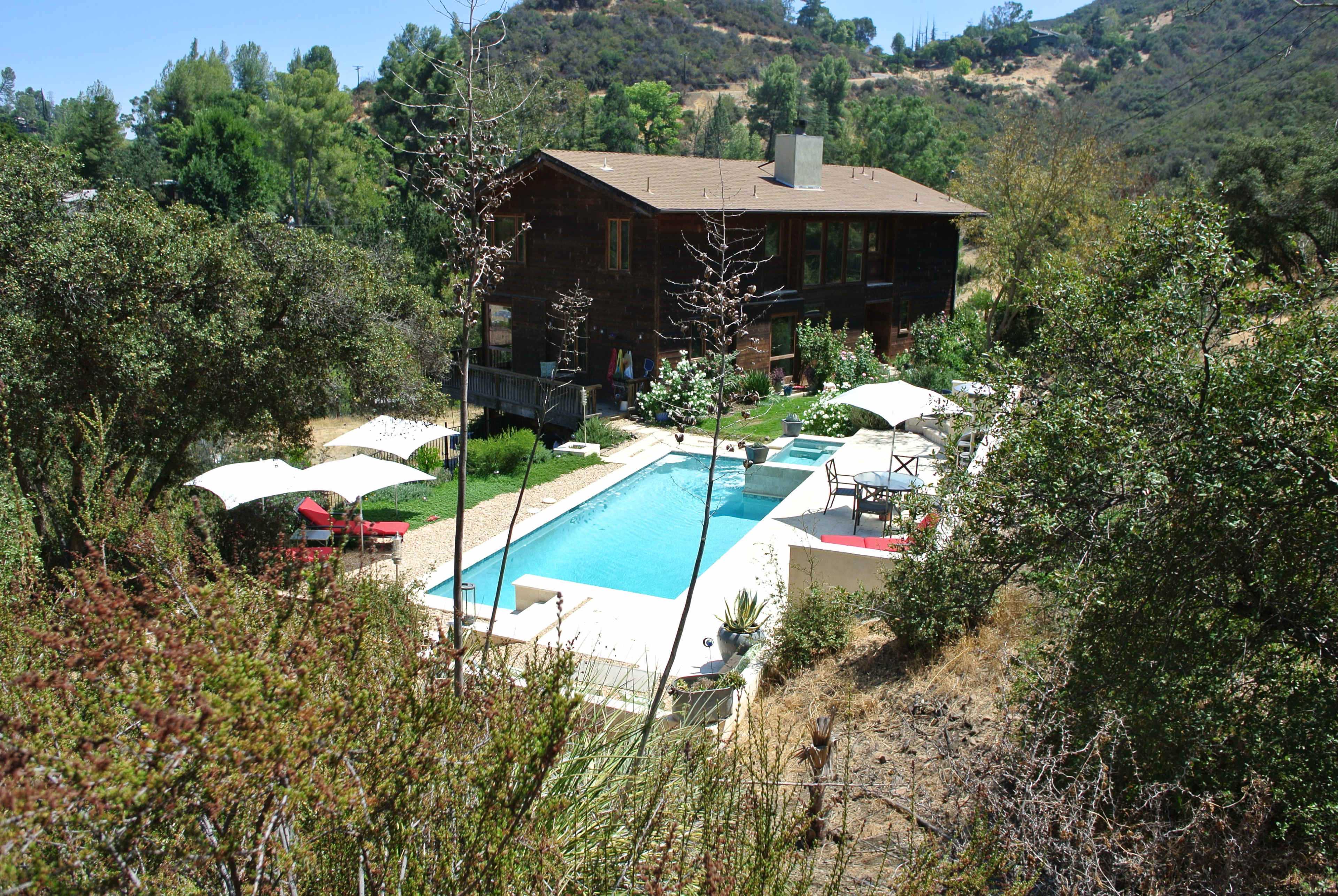 A modern house with large windows overlooks a rectangular swimming pool surrounded by lounge chairs and umbrellas, situated in a hilly landscape with greenery.