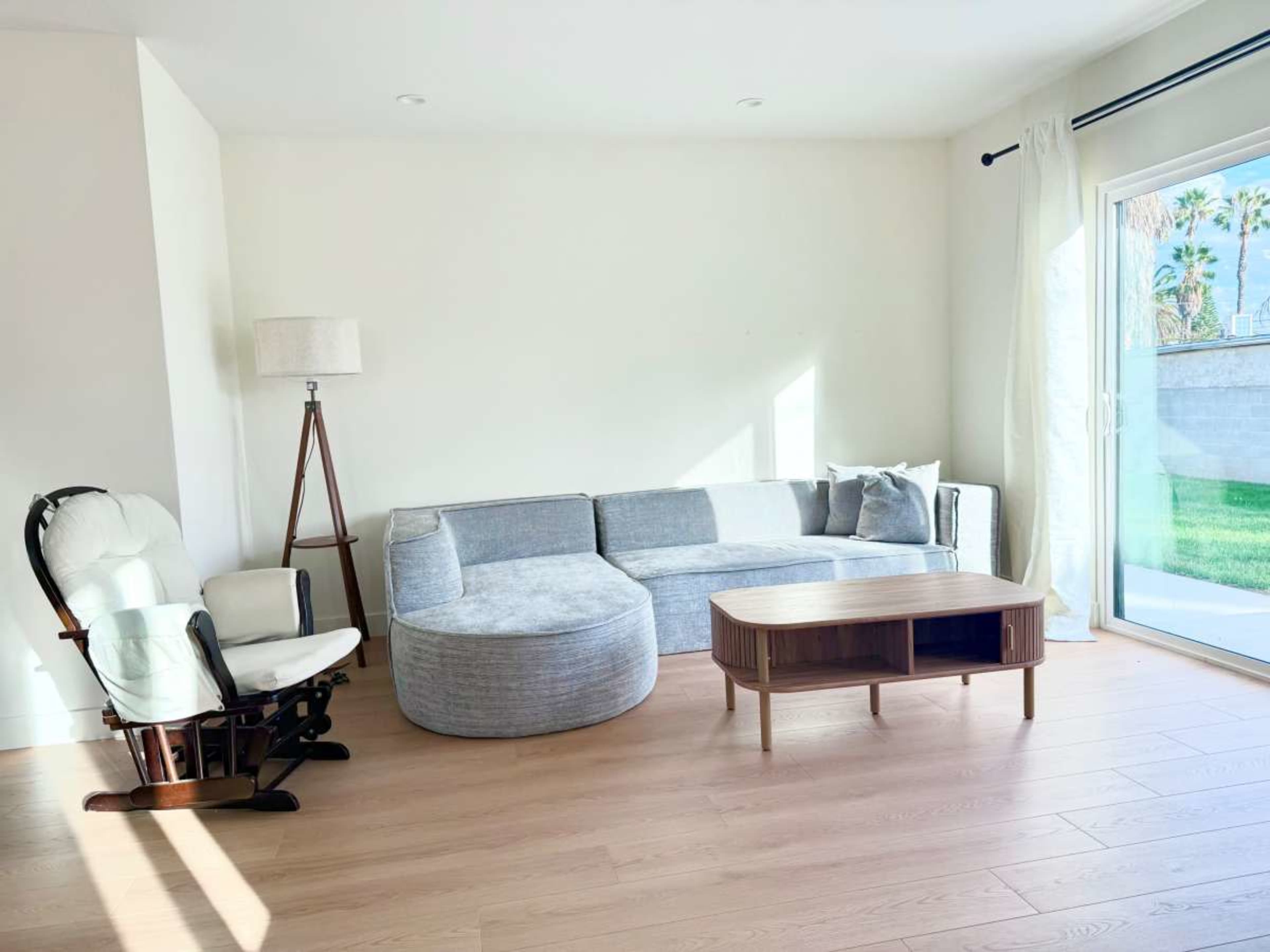 The image shows a modern living room with a gray sectional sofa, a round ottoman, a wooden coffee table, and a white rocking chair beside a large window.