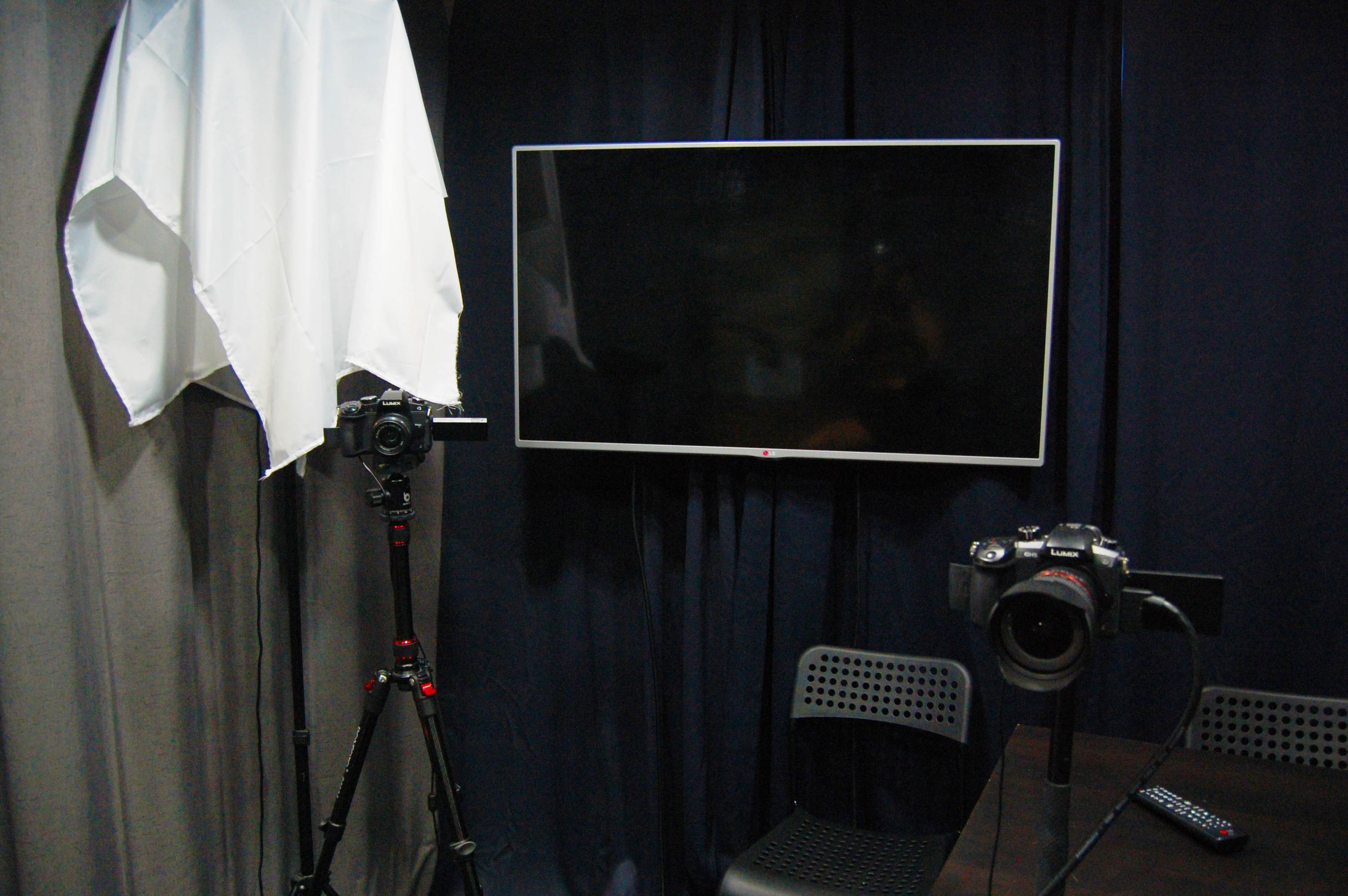 The image shows a photography setup with a camera on a tripod, a large television screen, and a second camera positioned on a table.