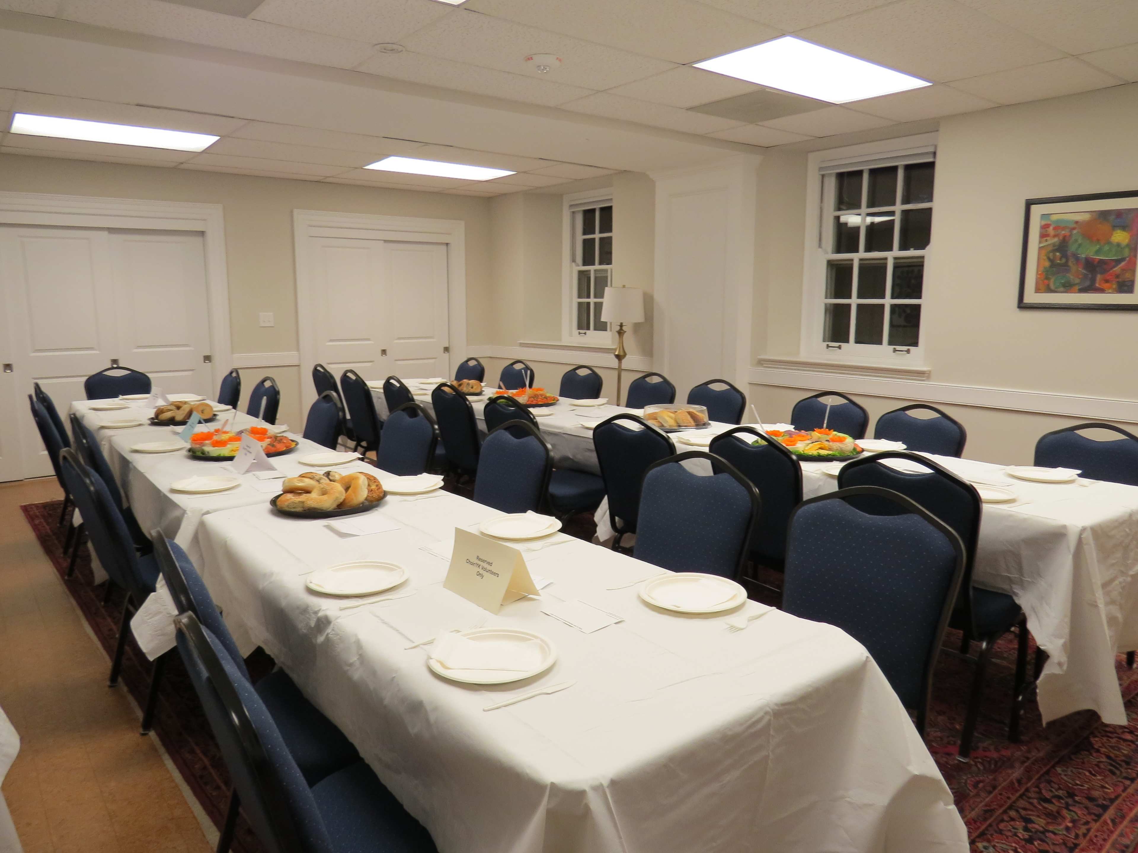 A dining room features several long tables set with white tablecloths and place settings, surrounded by blue chairs, and adorned with trays of food.