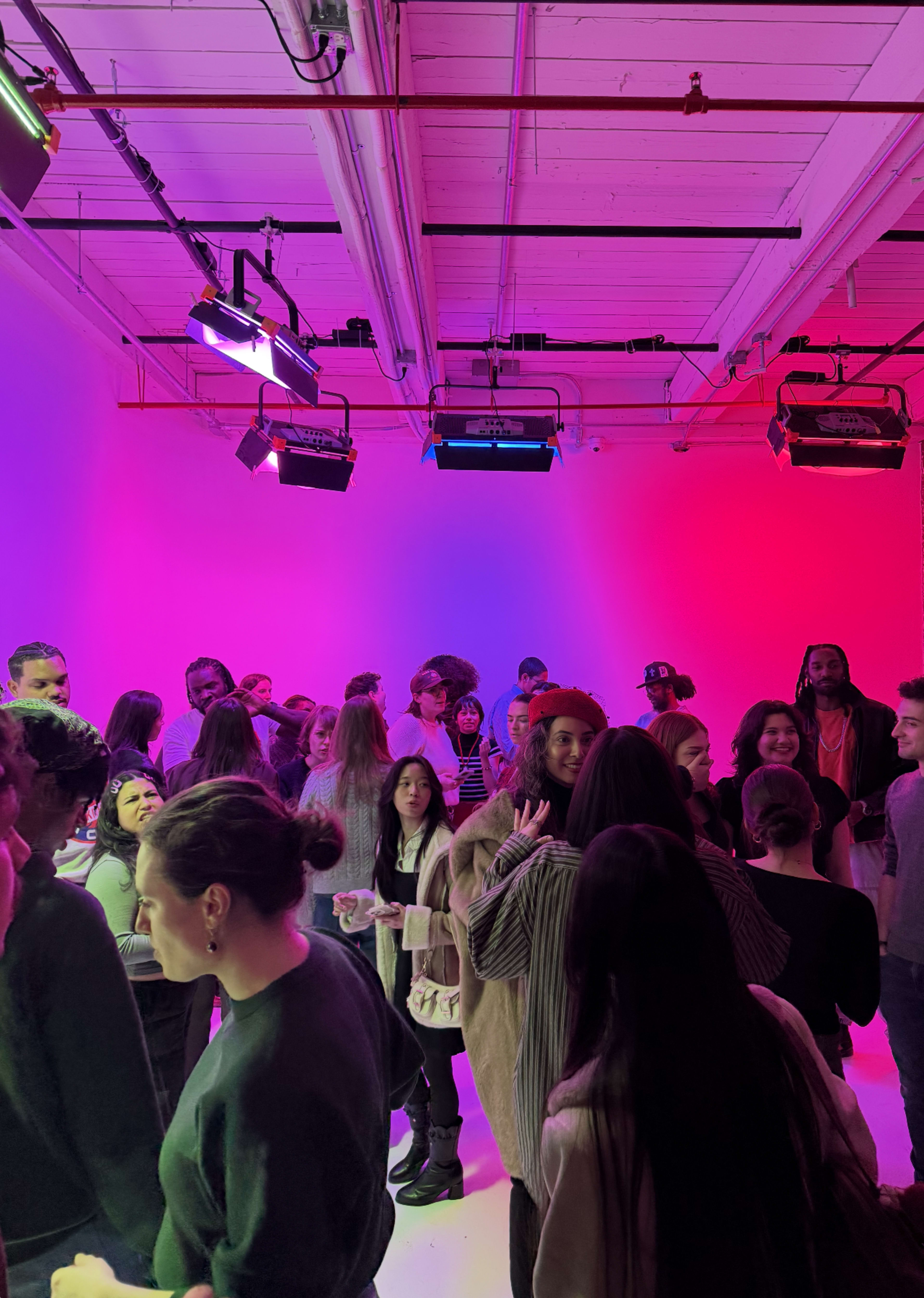 A large group of people is mingling in a brightly lit space with pink and purple backgrounds.
