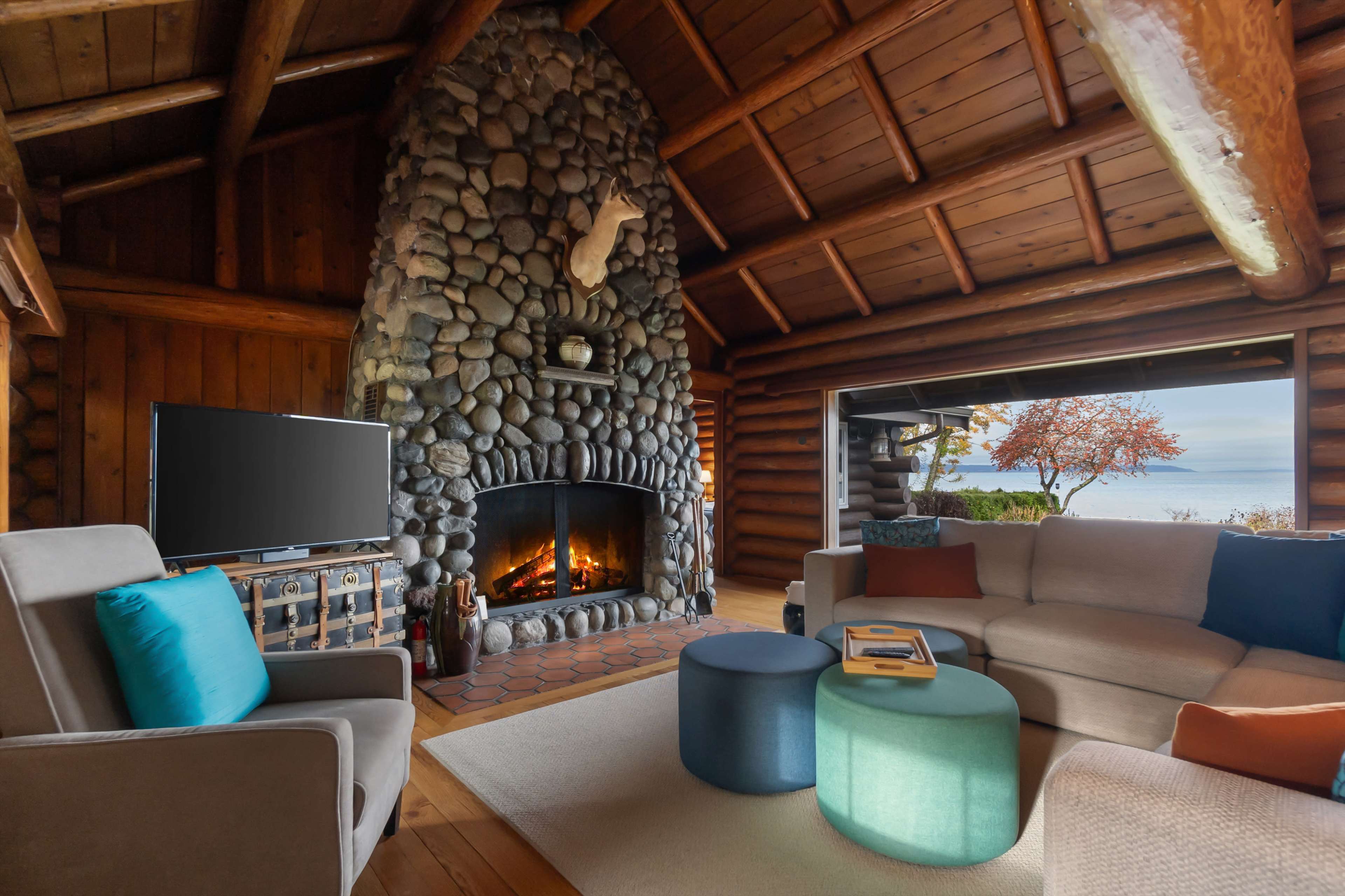 The image showcases a cozy interior of a log cabin featuring a large stone fireplace, comfortable seating, and a view of the outdoors through large windows.