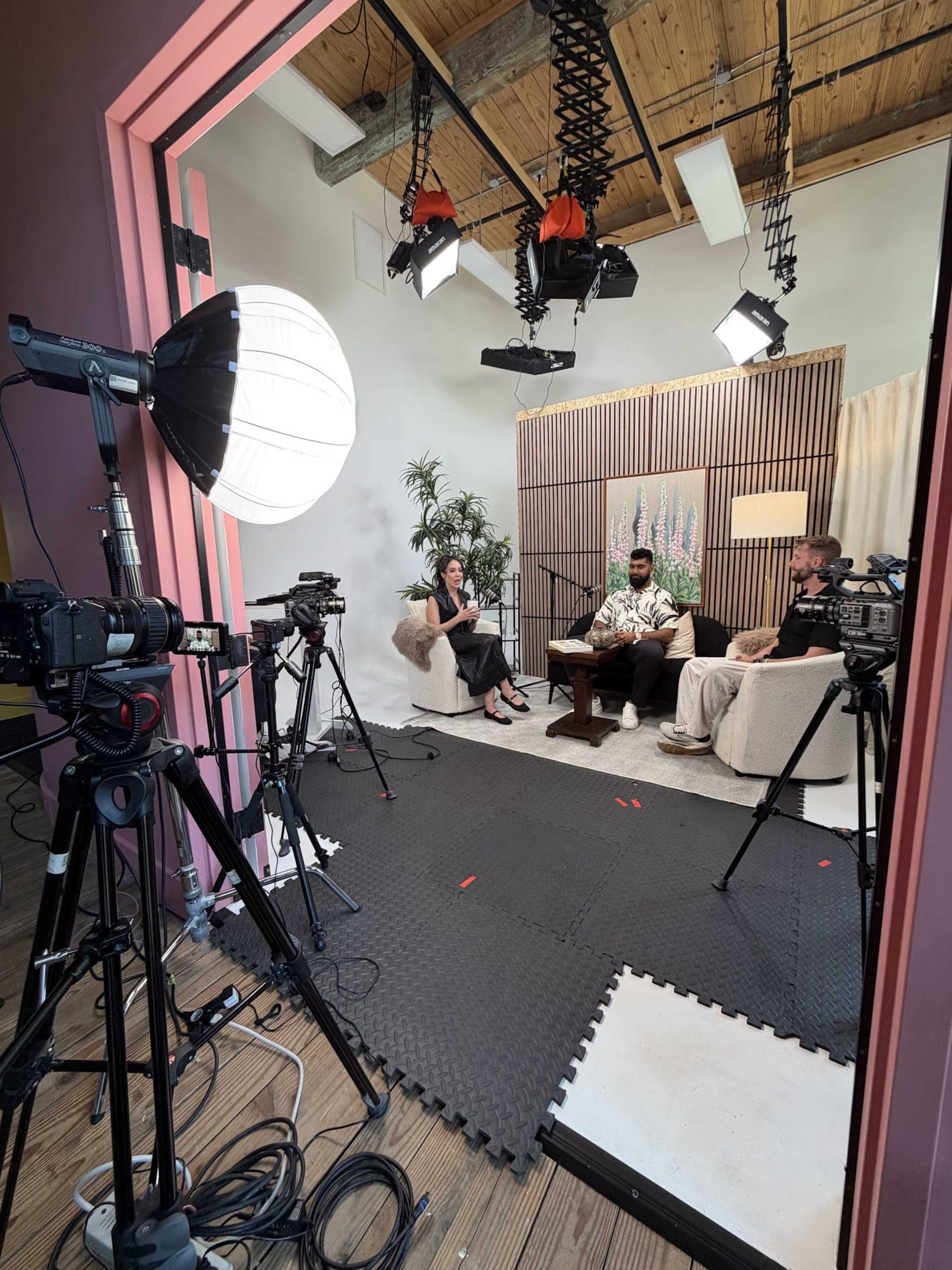 A well-lit studio setup features three people seated and discussing, with multiple cameras and lighting equipment arranged around them.