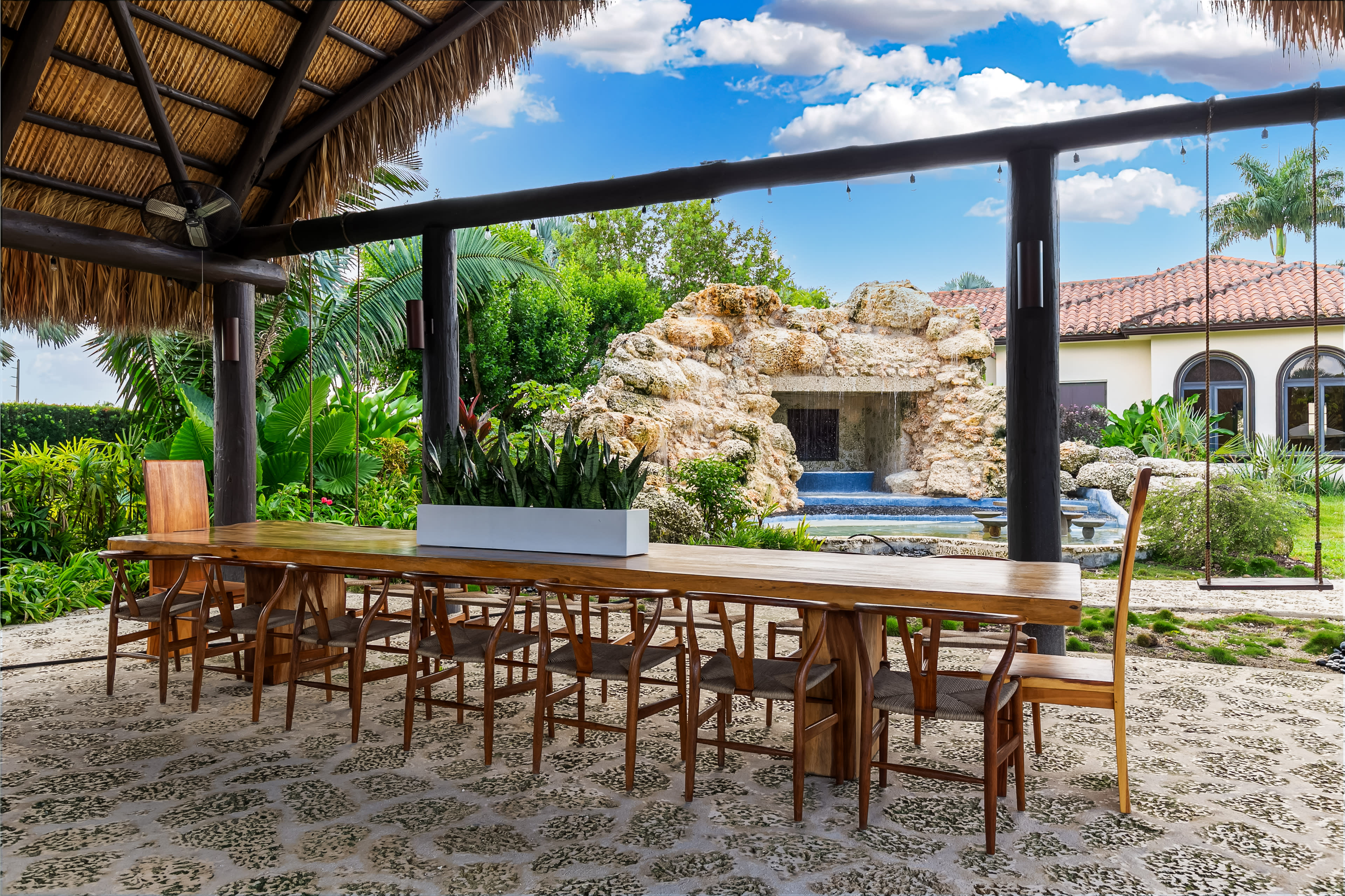 A long wooden dining table with chairs is set under a thatched roof, overlooking a landscaped garden with a rock fountain.