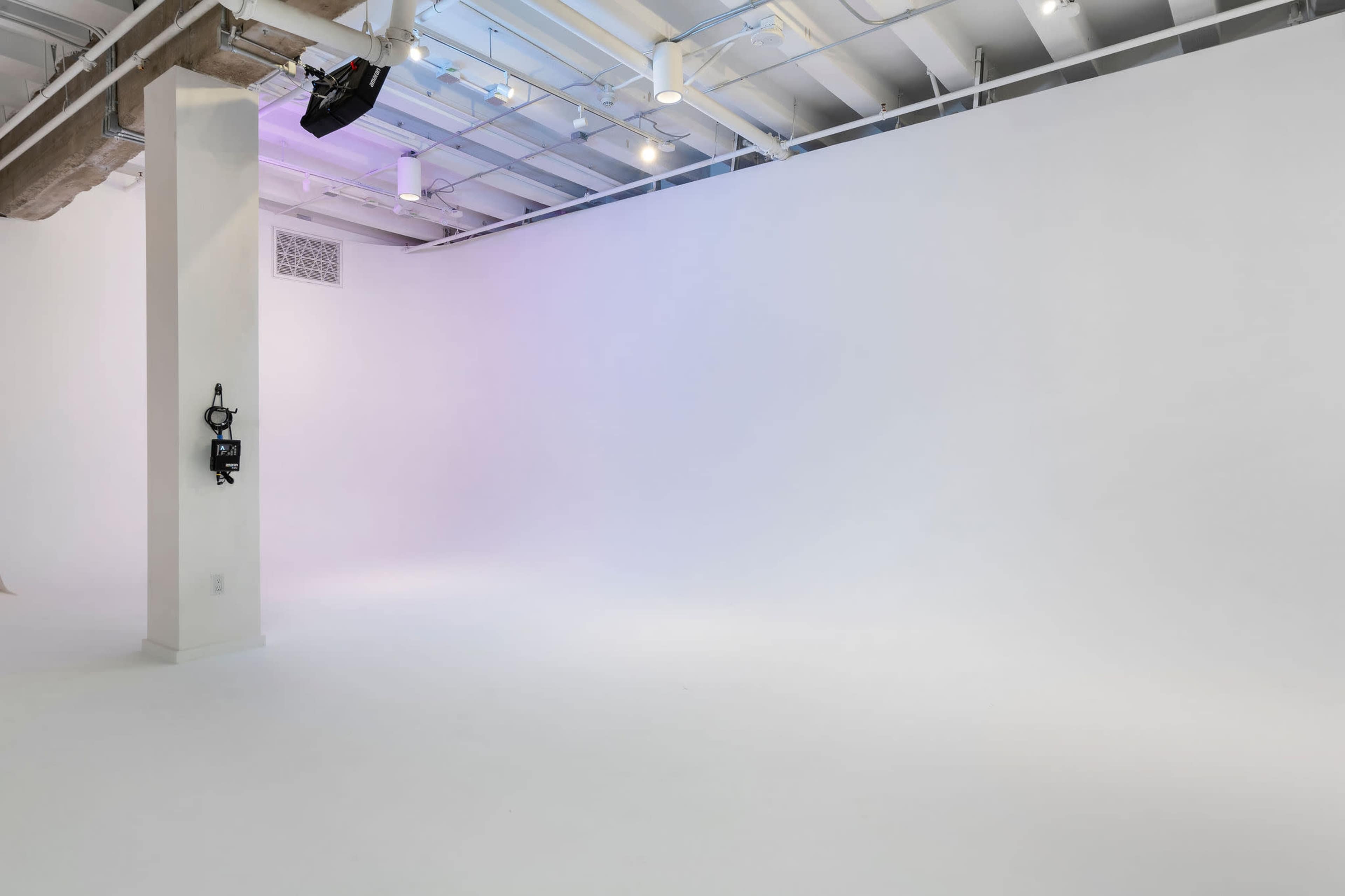 A spacious, empty studio with white walls and flooring, featuring overhead lighting and a visible support column.