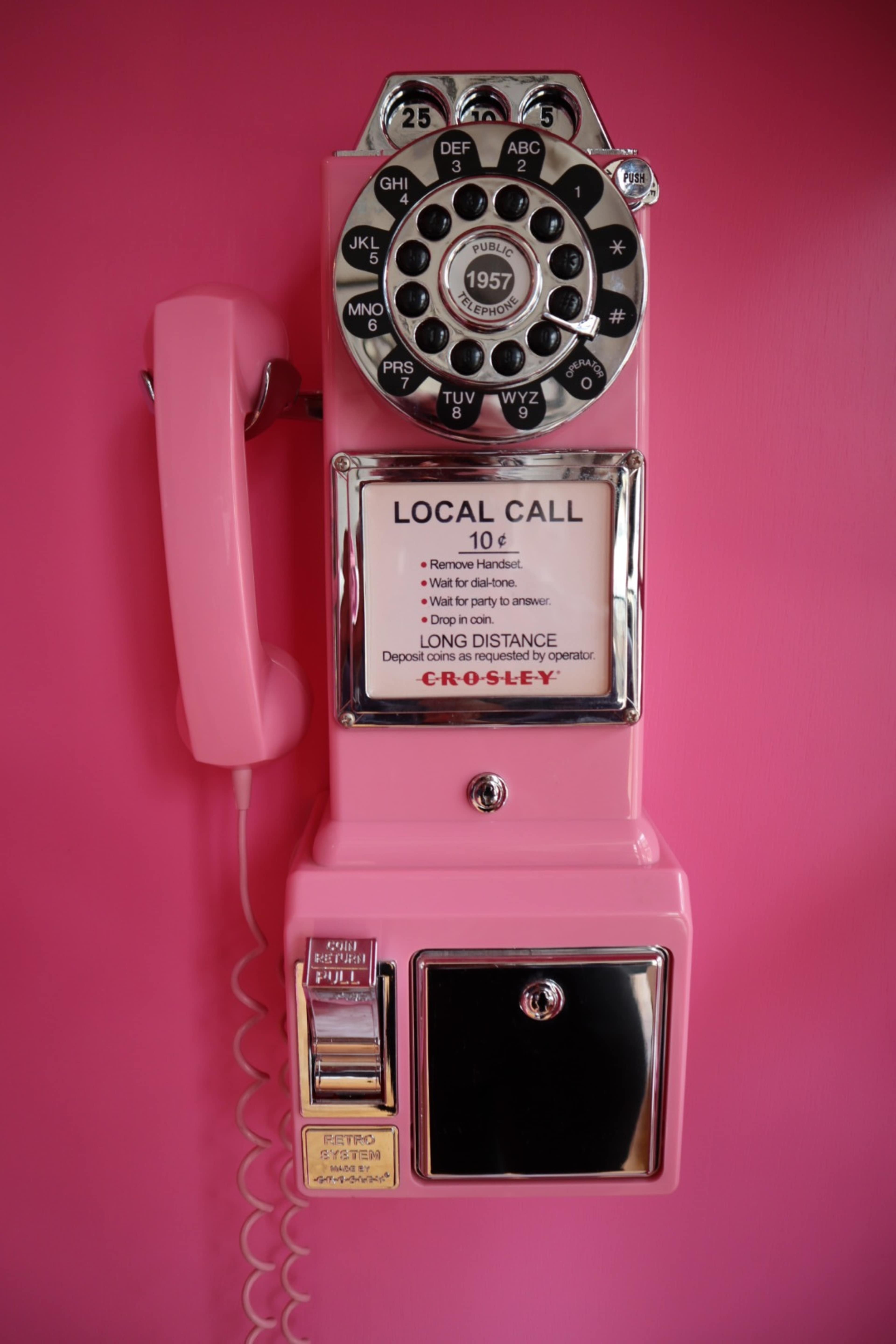 A vintage pink rotary phone with a coin slot is mounted on a bright pink wall.