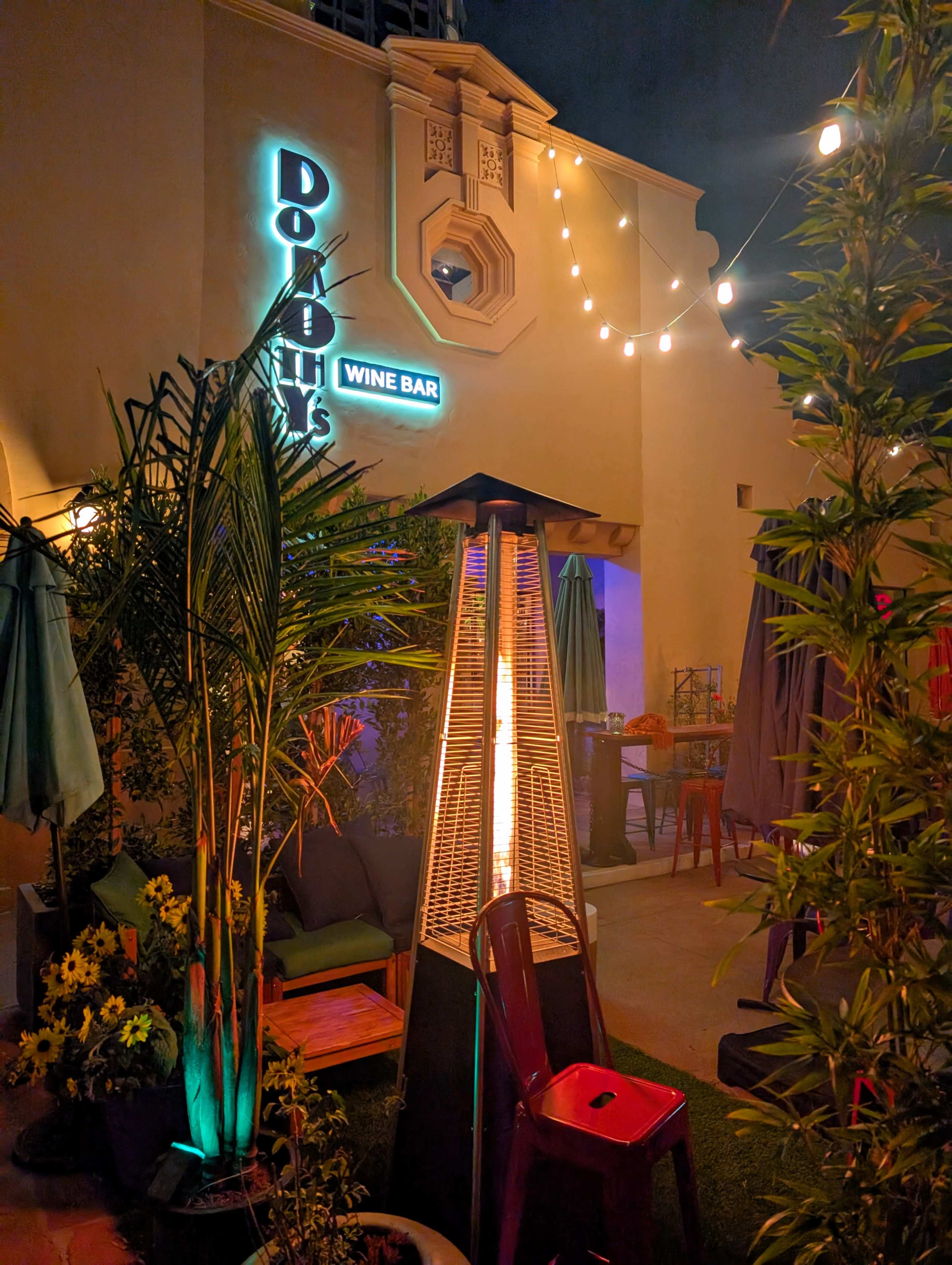 The image shows an outdoor wine bar area at night, featuring a tall patio heater surrounded by plants and illuminated by string lights.