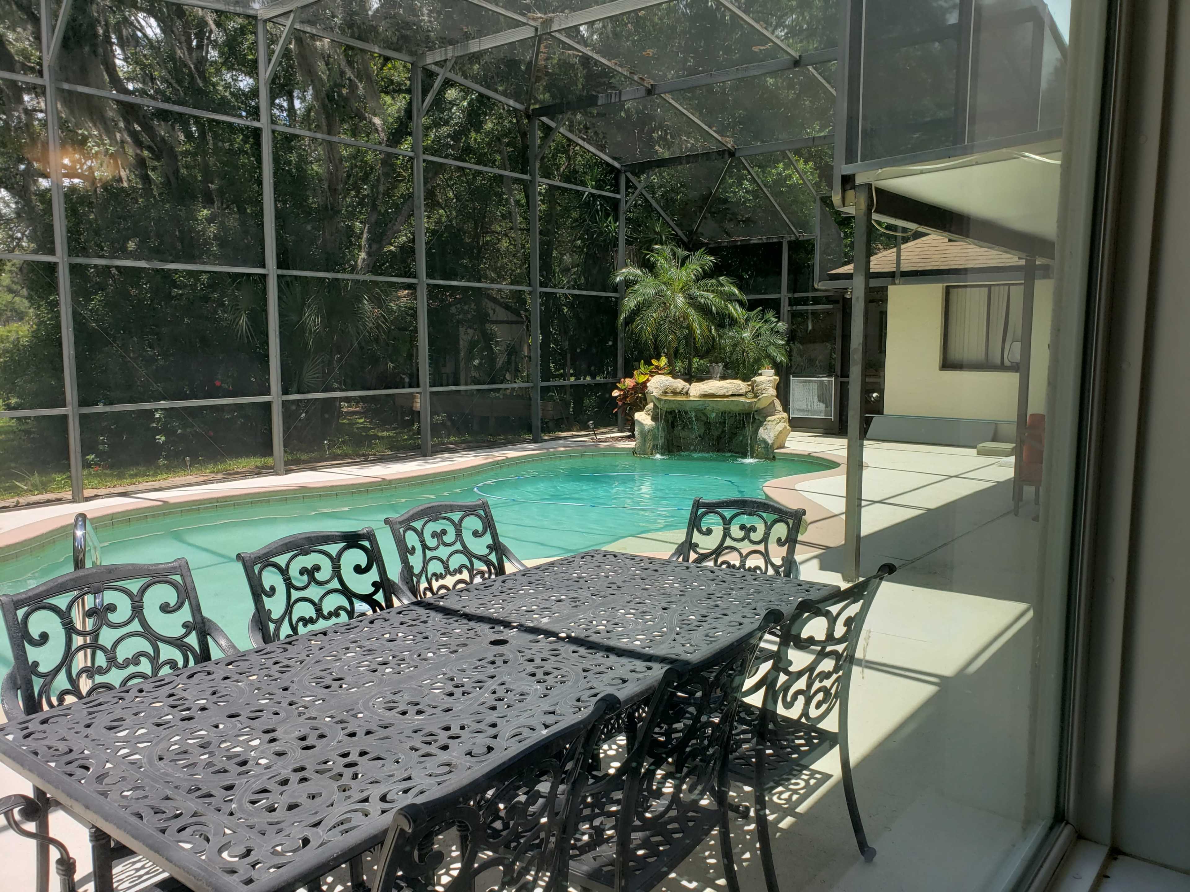 A screen-enclosed patio features a table and chairs beside a swimming pool with a waterfall and tropical plants.