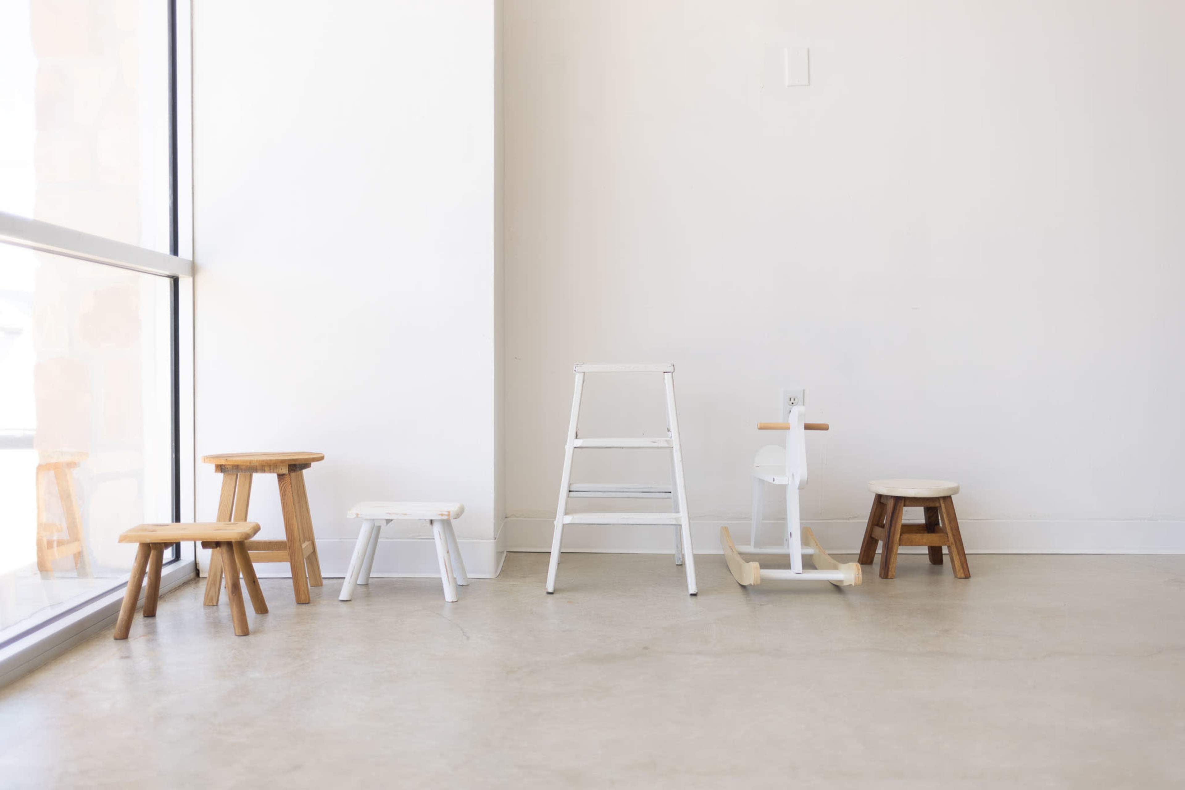 The image shows a minimalist interior with five various wooden stools and a white rocking chair arranged near a large window.